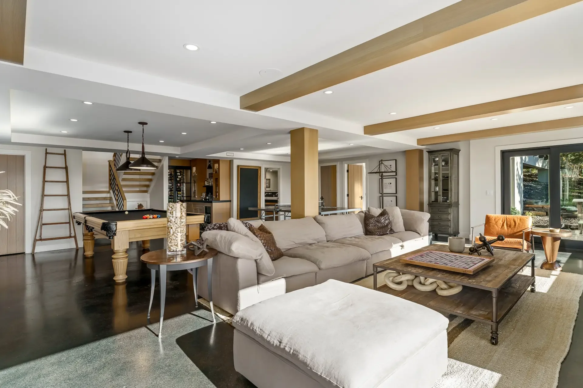 Open-concept living room with beige sectional sofa, wooden coffee table, and a pool table in the background. Large sliding glass doors reveal an outdoor area with greenery. The ceiling has exposed wood beams, and there are various decorative and functional items throughout the space.