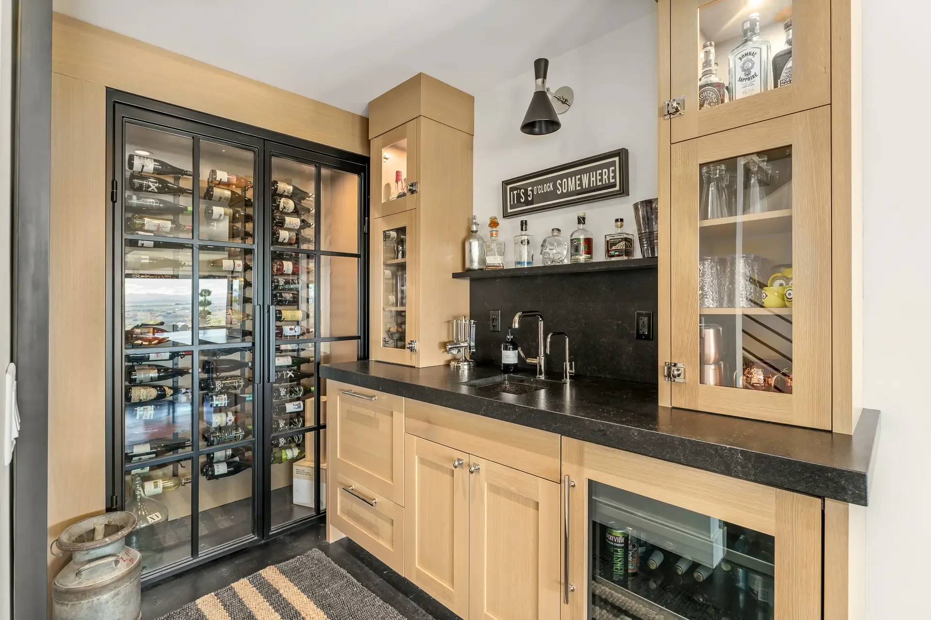 A home bar area with a black wine fridge, glass cabinet with liquor bottles, granite countertop, and natural wood cabinets.