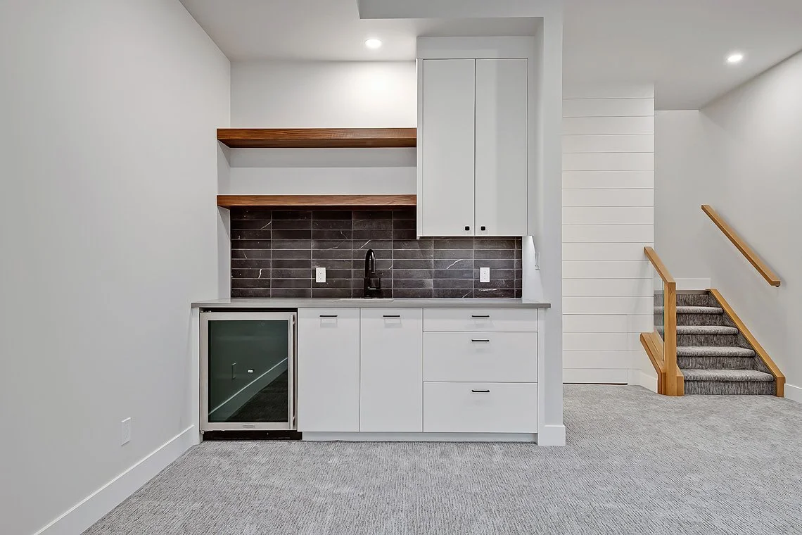 Small kitchenette with white cabinets, black faucet, dark tile backsplash, mini fridge, open shelves, and staircase with wooden handrails.
