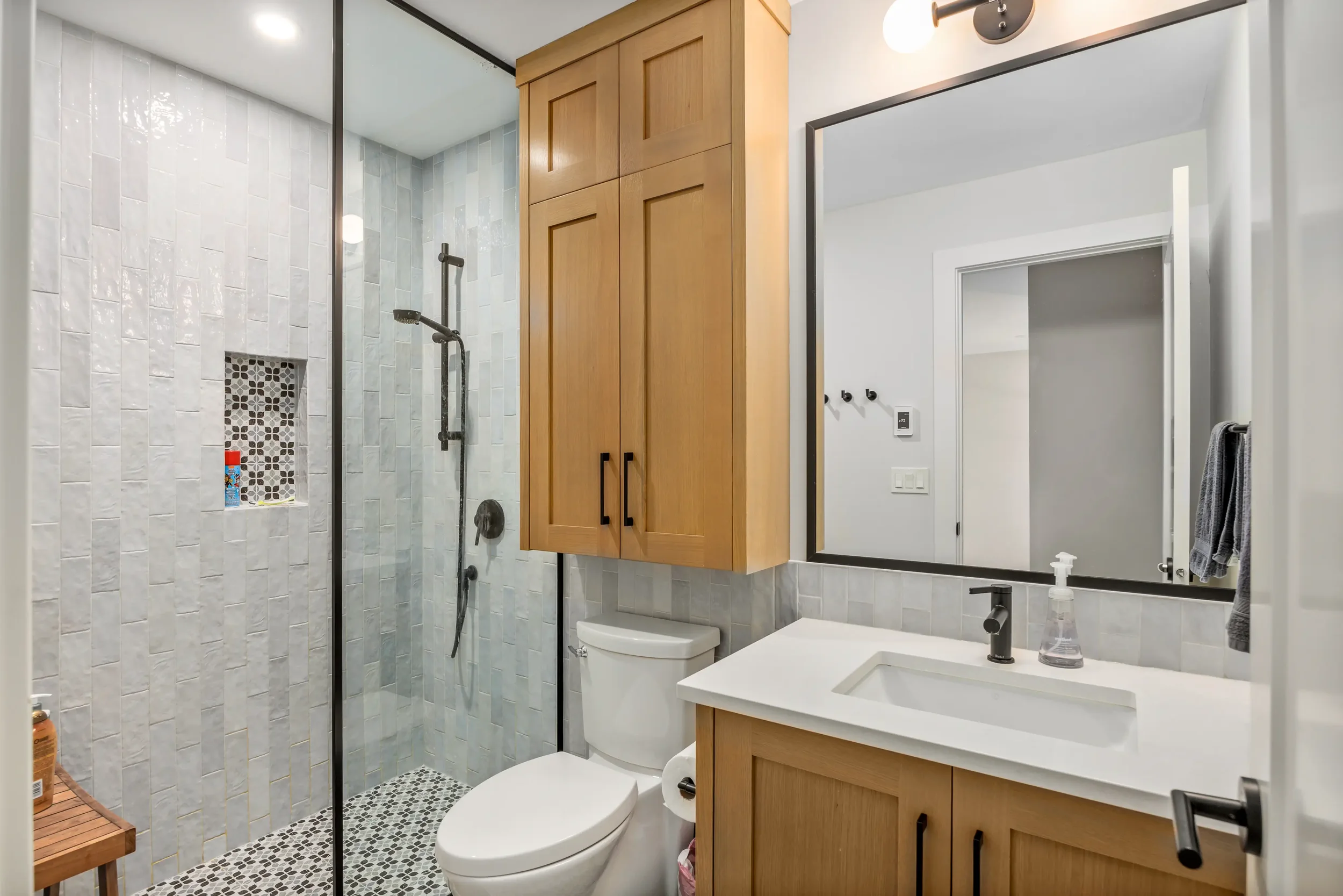 A modern bathroom featuring a shower with white tiles and a black showerhead, a wooden cabinet above the toilet, a large mirror, a white sink with a black faucet, and a towel hanging on the wall.