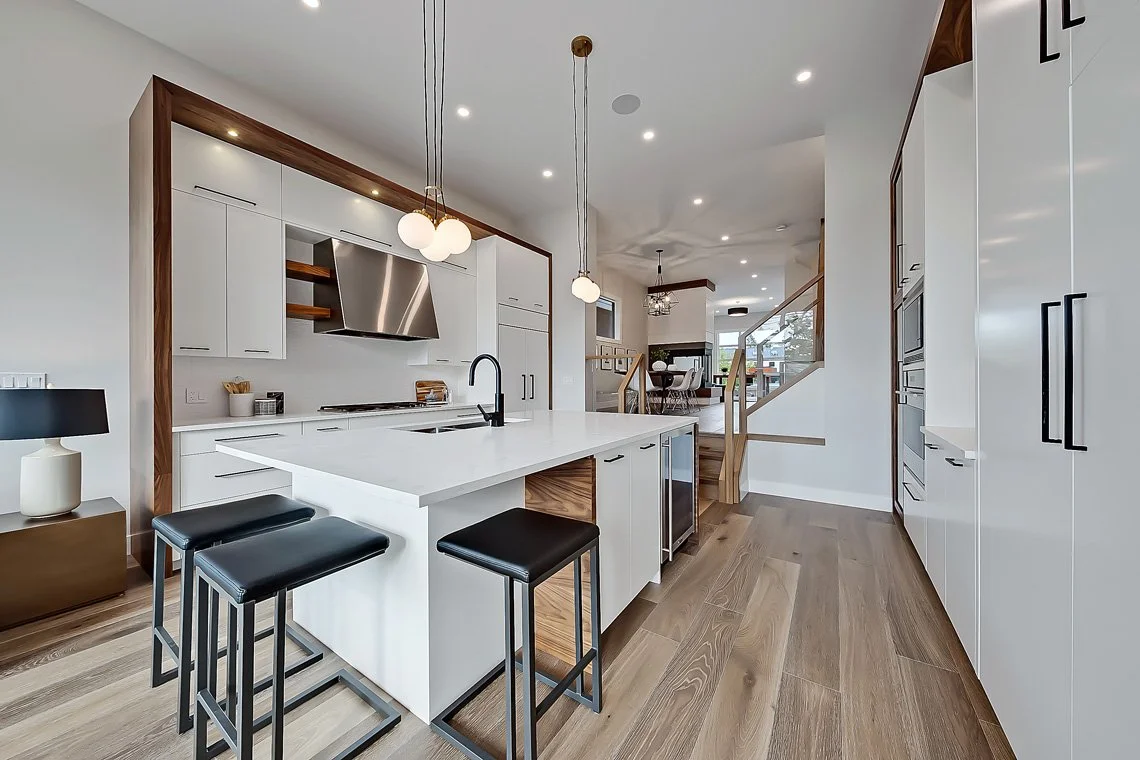 Modern open-concept kitchen with white cabinets, a central island with black stools, wooden accents, pendant lights, and a view into the dining and living areas.