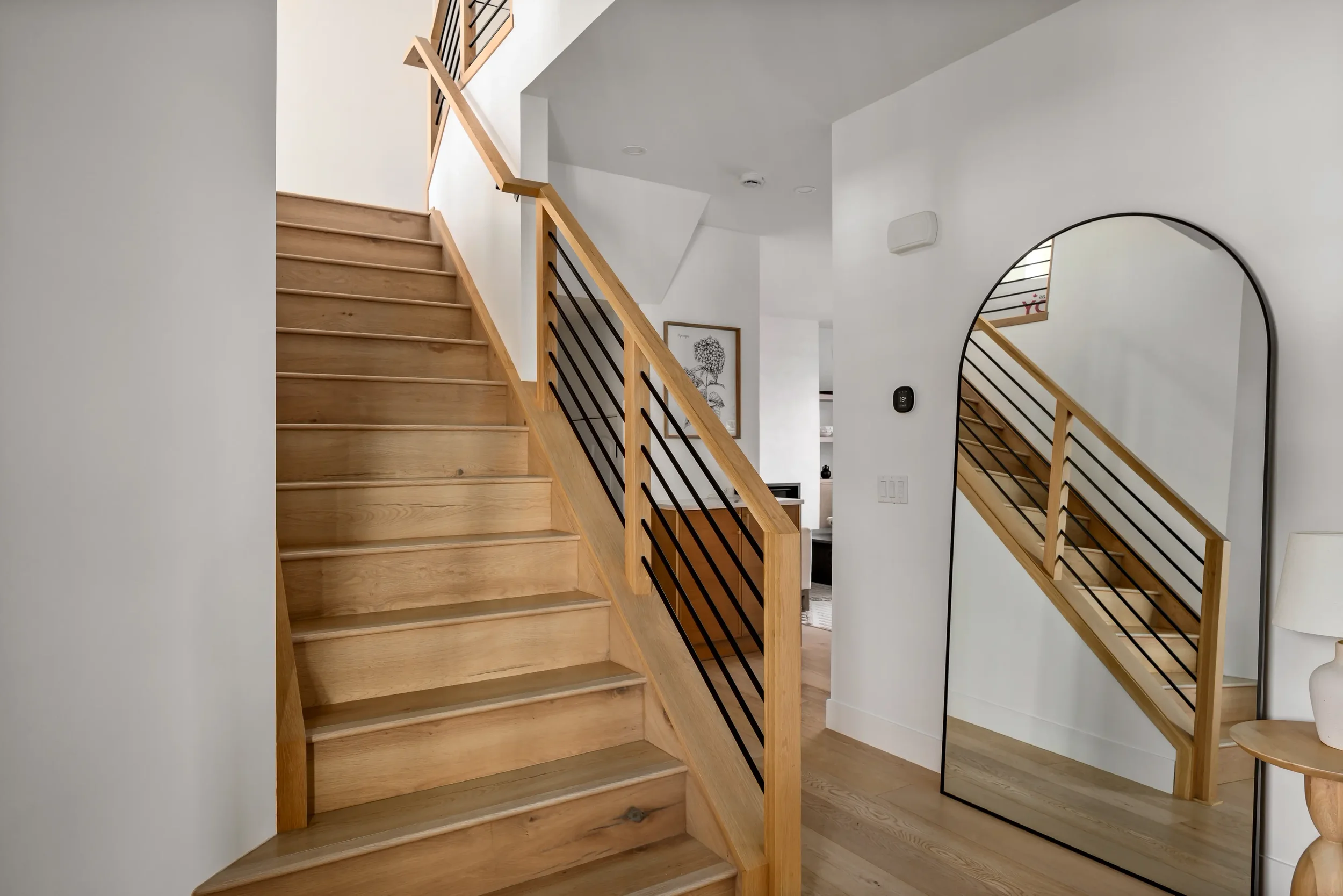 Interior of a modern house with a wooden staircase, a large mirror with a black frame, and minimalistic decor including a framed line drawing and a white side table.
