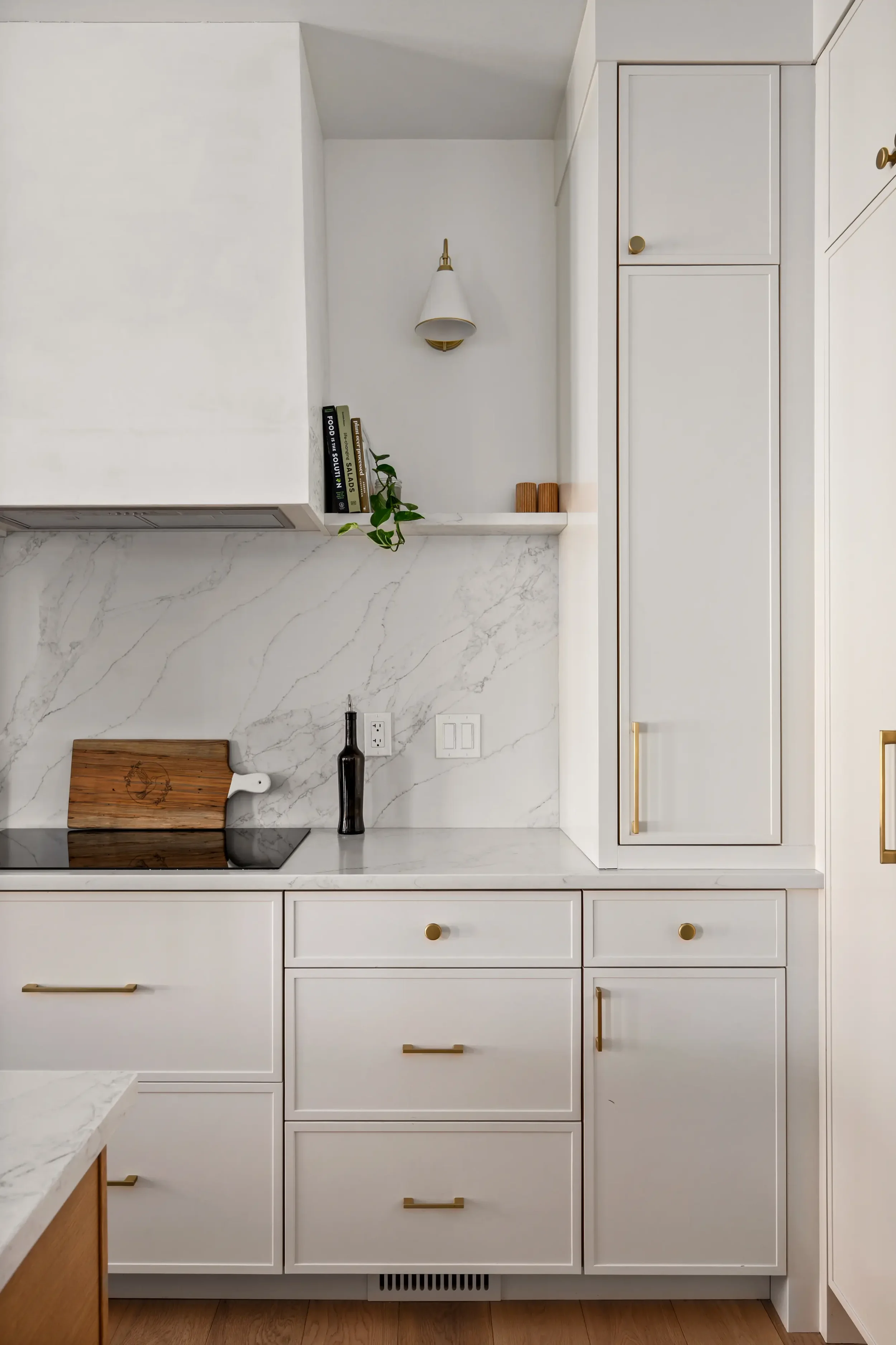 White kitchen cabinets with gold handles, a marble countertop, and a cutting board with a bottle of oil. Small shelf with books, a plant, and a wall-mounted sconce.