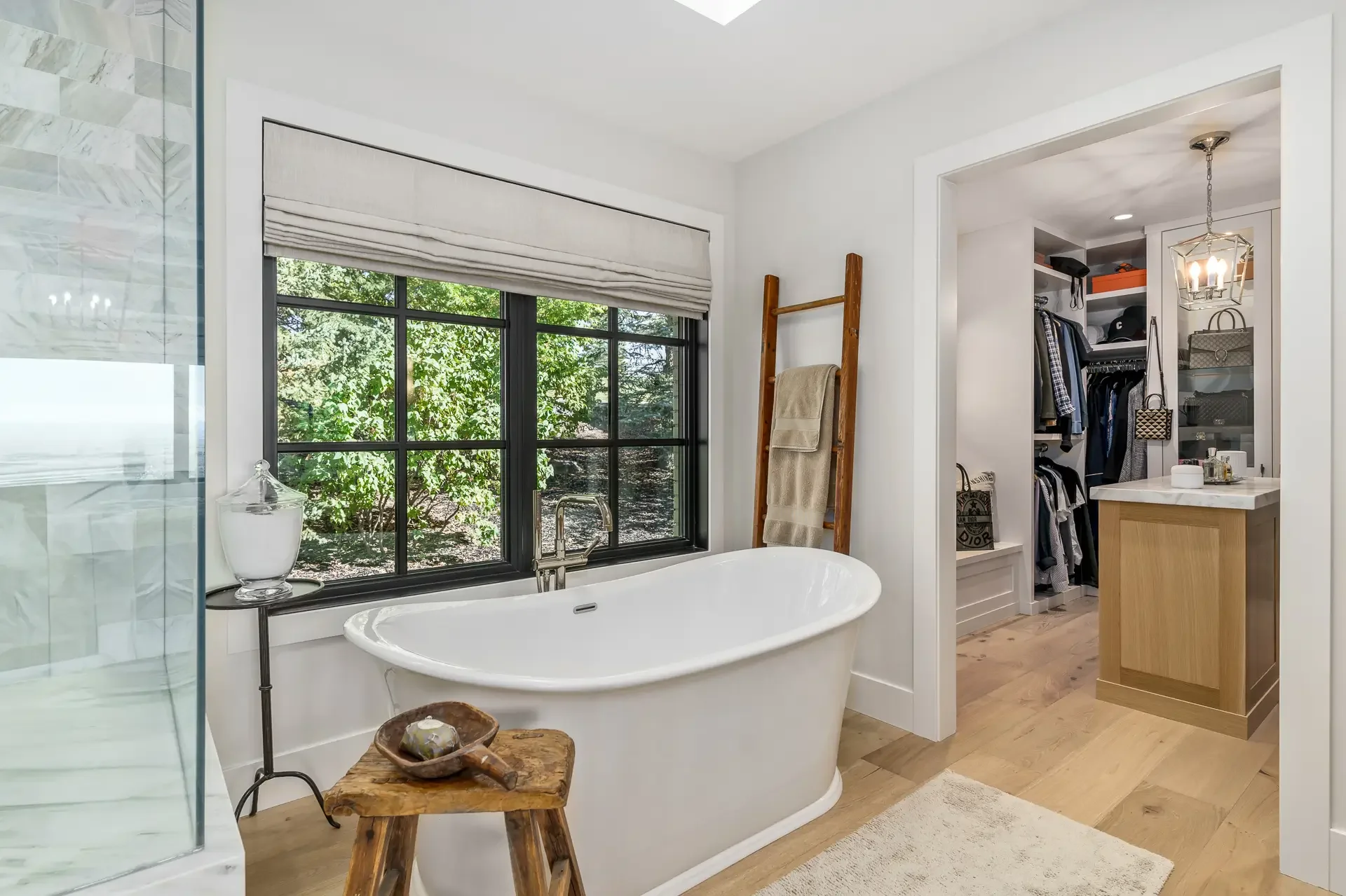Modern bathroom with freestanding bathtub, window view of greenery, wooden stool, and walk-in closet with hanging clothes and accessories.