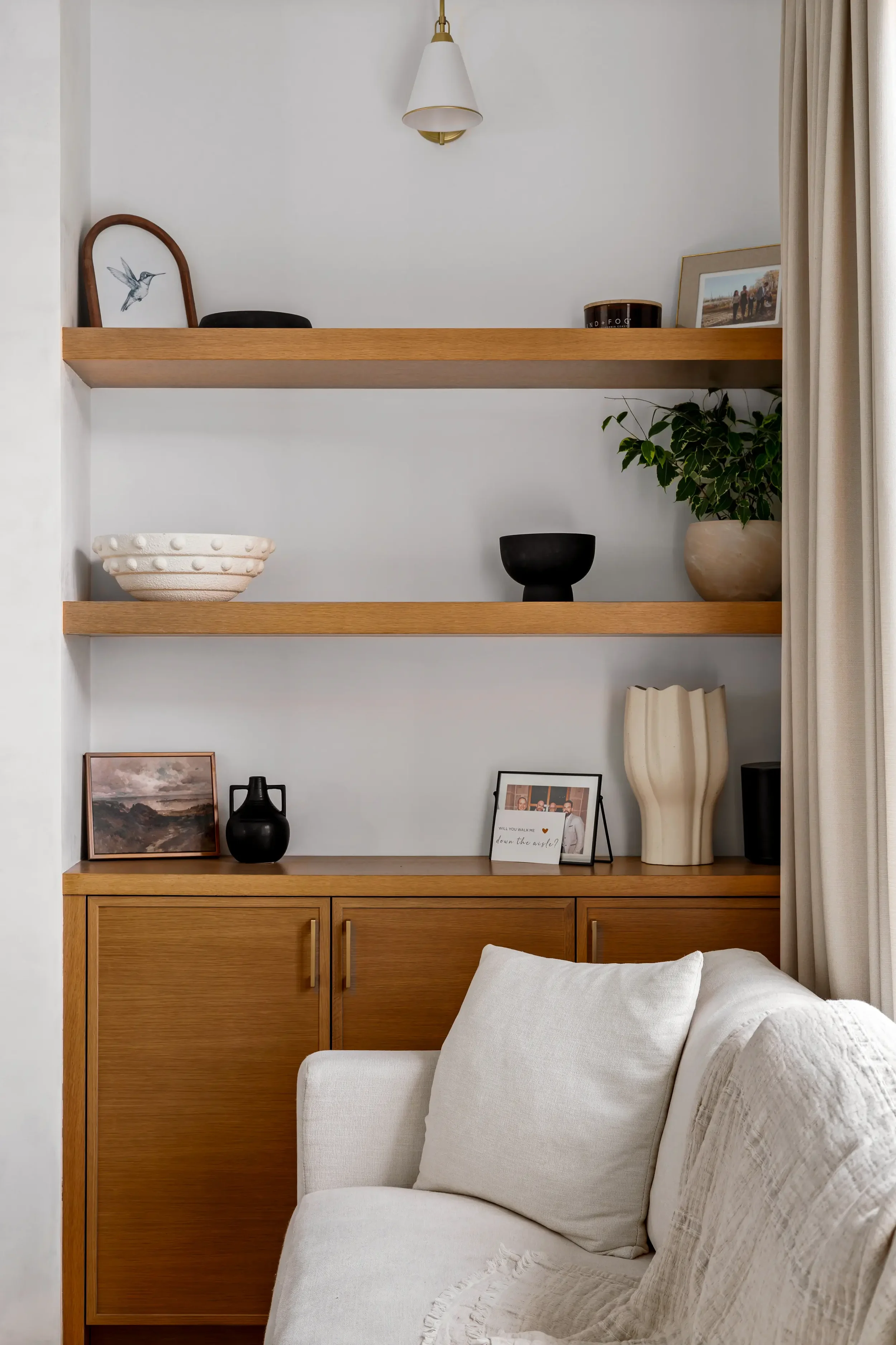 Living room with wooden shelving displaying framed artwork, photographs, decorative bowls, potted plant, pitcher, and various decor items, with part of a white sofa and curtain visible.