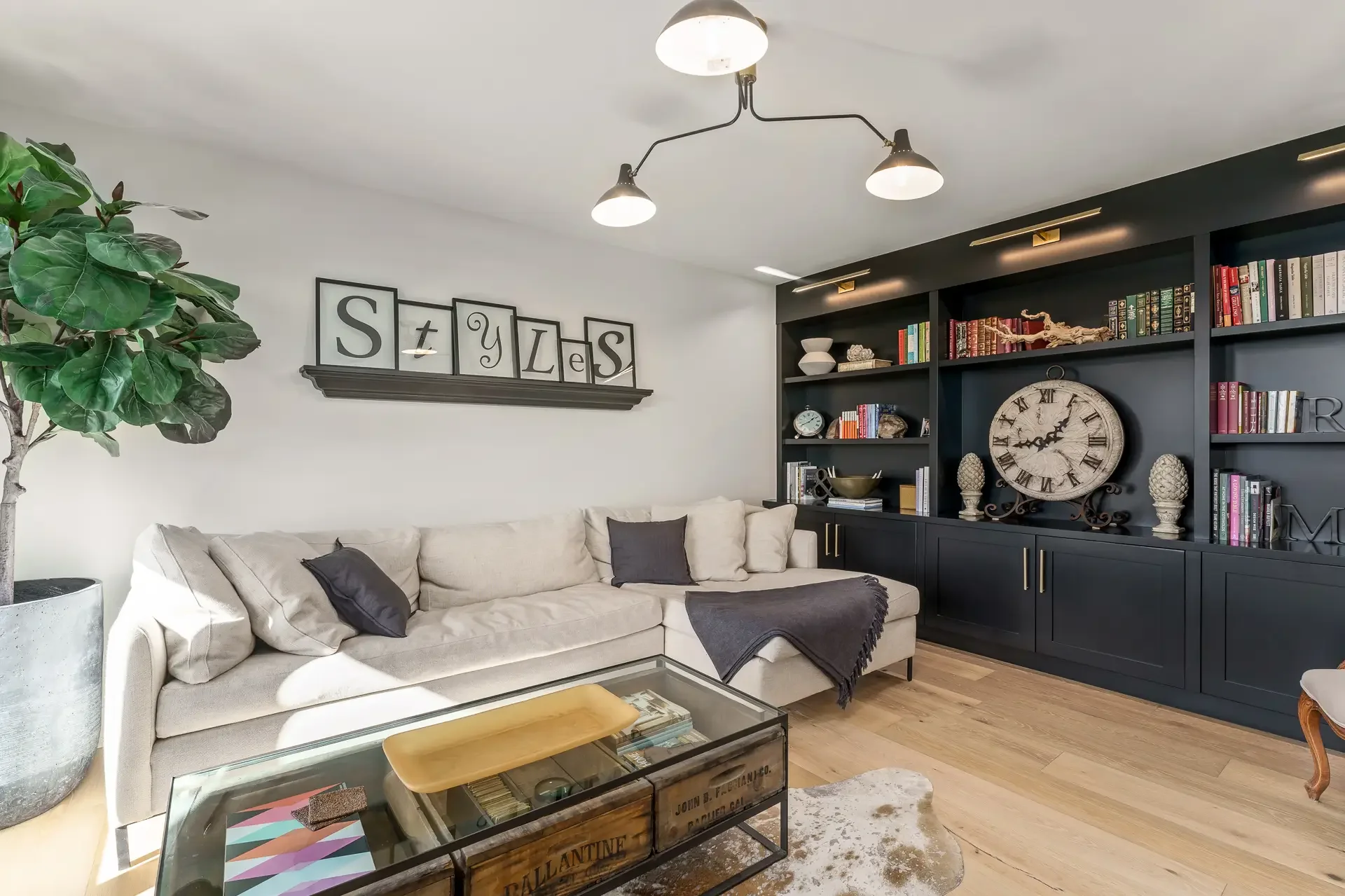 Living room with a beige sofa, dark wall bookshelf with books and decorative items, large clock, and potted plant, modern ceiling lights, and wooden flooring.