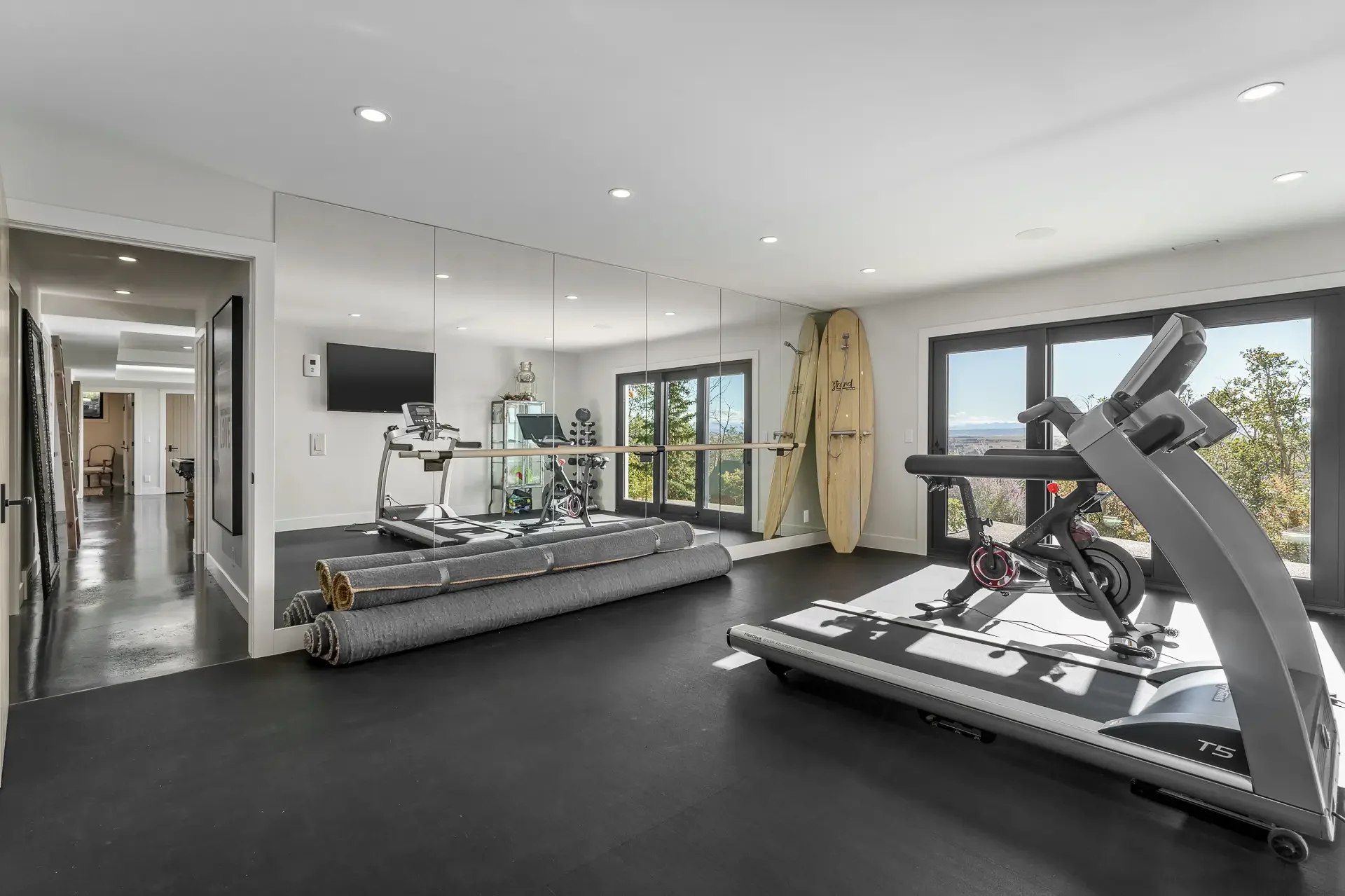 Modern home gym with treadmill, stationary bike, and surfboards near large windows showing trees and mountains outside.