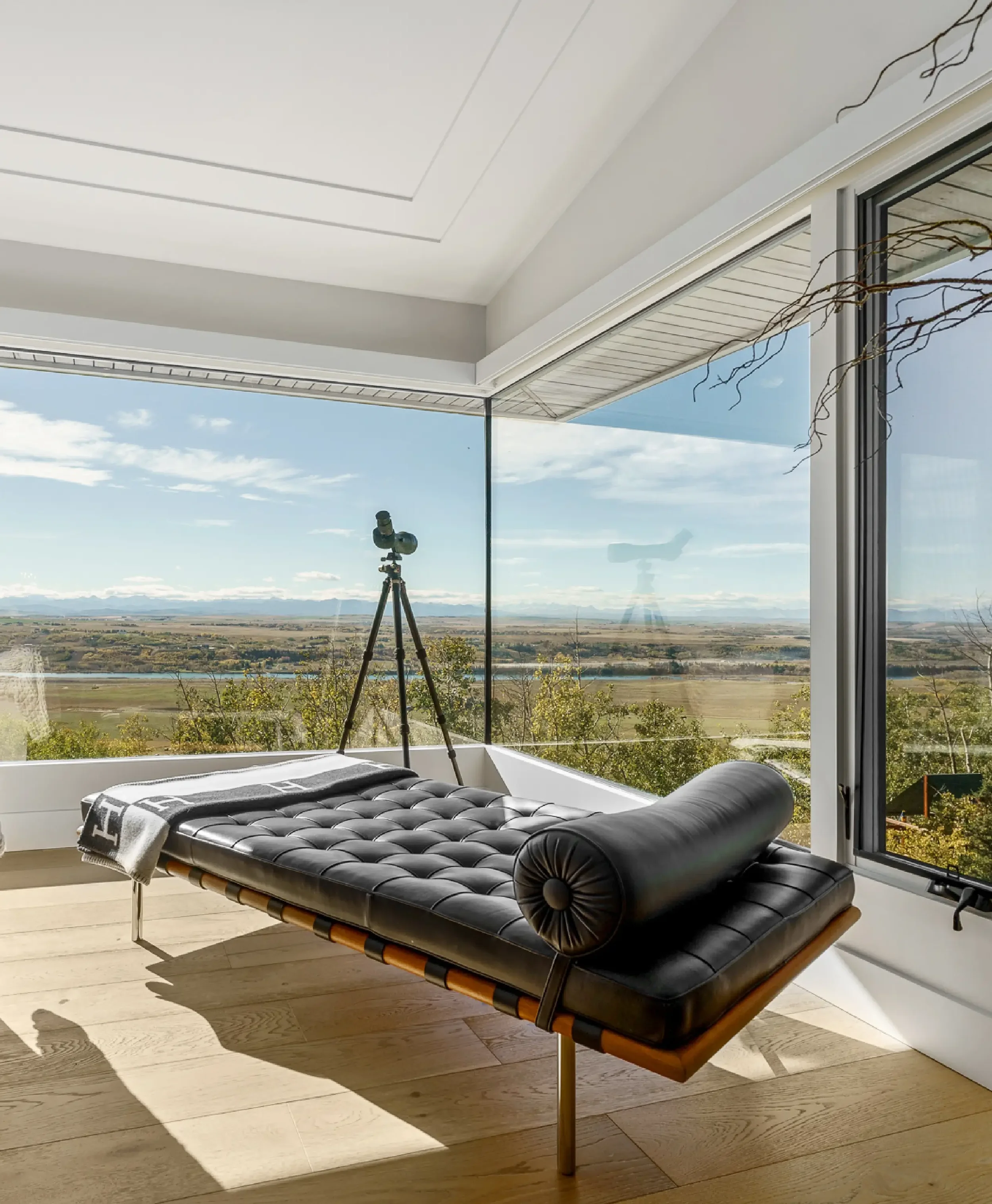 A modern black leather lounge chair with a wooden frame and a cylindrical rolled pillow at one end, placed near a large window showing an outdoor landscape with sky, clouds, and distant fields, with a tripod-mounted telescope or binoculars inside the room.