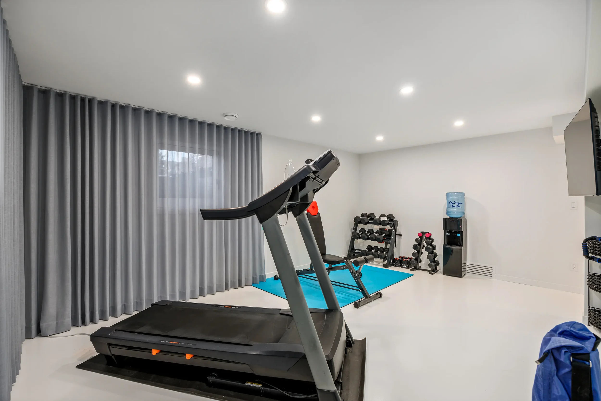 A home gym with a treadmill, dumbbells, a water cooler, and curtains in the background.