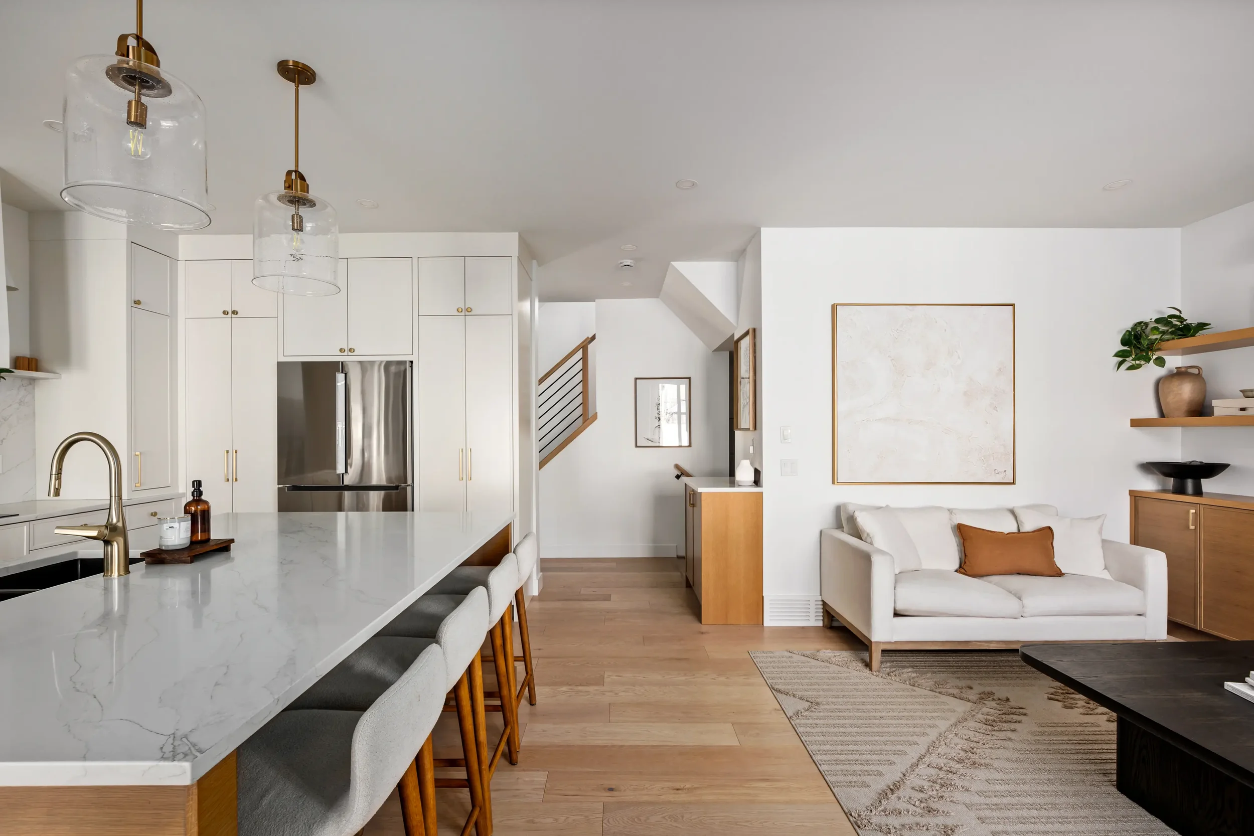 Open-concept living room and kitchen with white cabinets, marble island, and wooden furniture. White walls, a large abstract art piece, and minimalist decor. Modern lighting fixtures under the ceiling.
