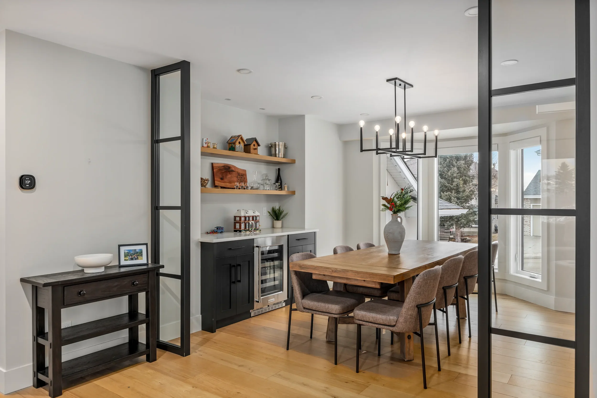 Modern dining area with wooden table, upholstered chairs, black and wood kitchen cabinets, open shelving with decor and glassware, large window, potted plant, and contemporary chandelier.