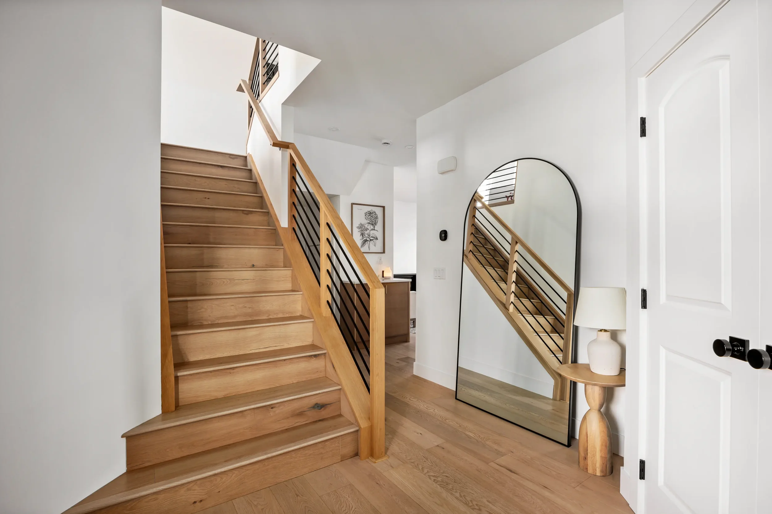 Interior view of a staircase with wooden steps and black metal railings, next to a white wall, a large mirror leaning against the wall, and a small side table with a white lamp and a ceramic vase.