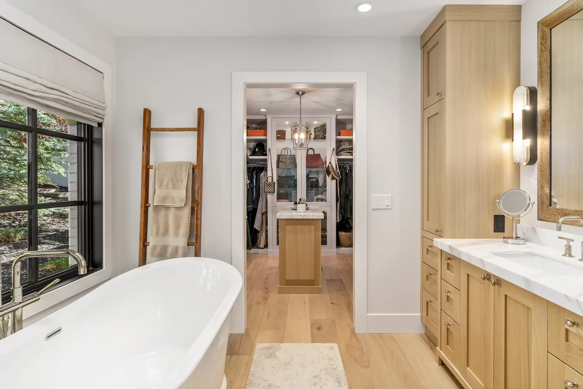 A bathroom with a white freestanding bathtub next to a window with a black frame and gray Roman shade. The room features light wood cabinets, a white marble countertop, and a wall-mounted light fixture. In the background, a walk-in closet with shelve