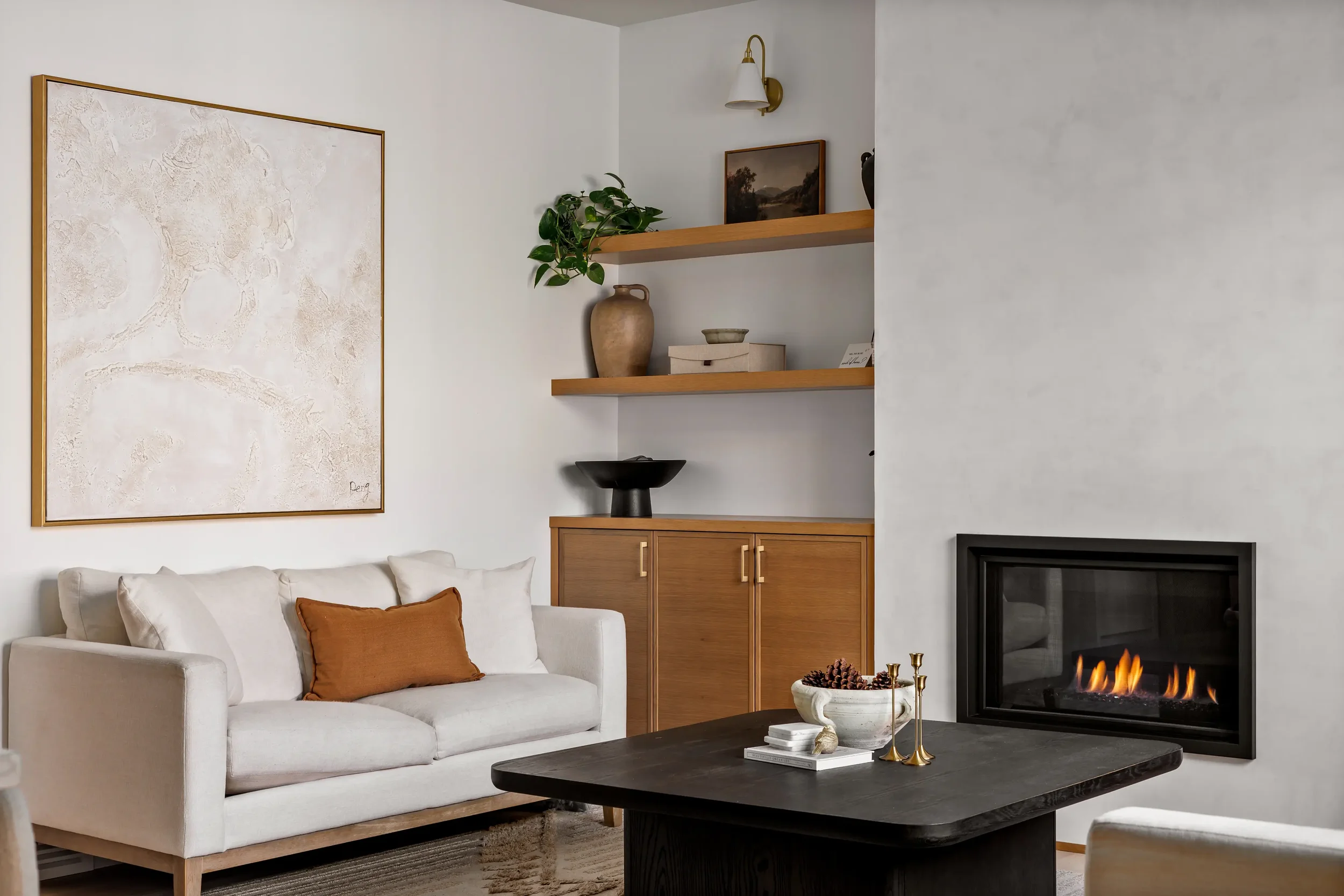 A cozy living room with a white sofa decorated with brown pillows, a black fireplace with visible flames, a dark coffee table with books and decorative items, wooden wall shelves with decor and a potted plant, a large textured wall art, and a white w