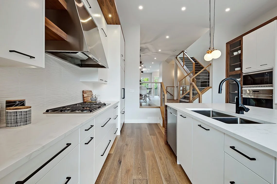 Modern kitchen with white cabinets, black handles, wooden flooring, and a staircase with wooden railing in the background.