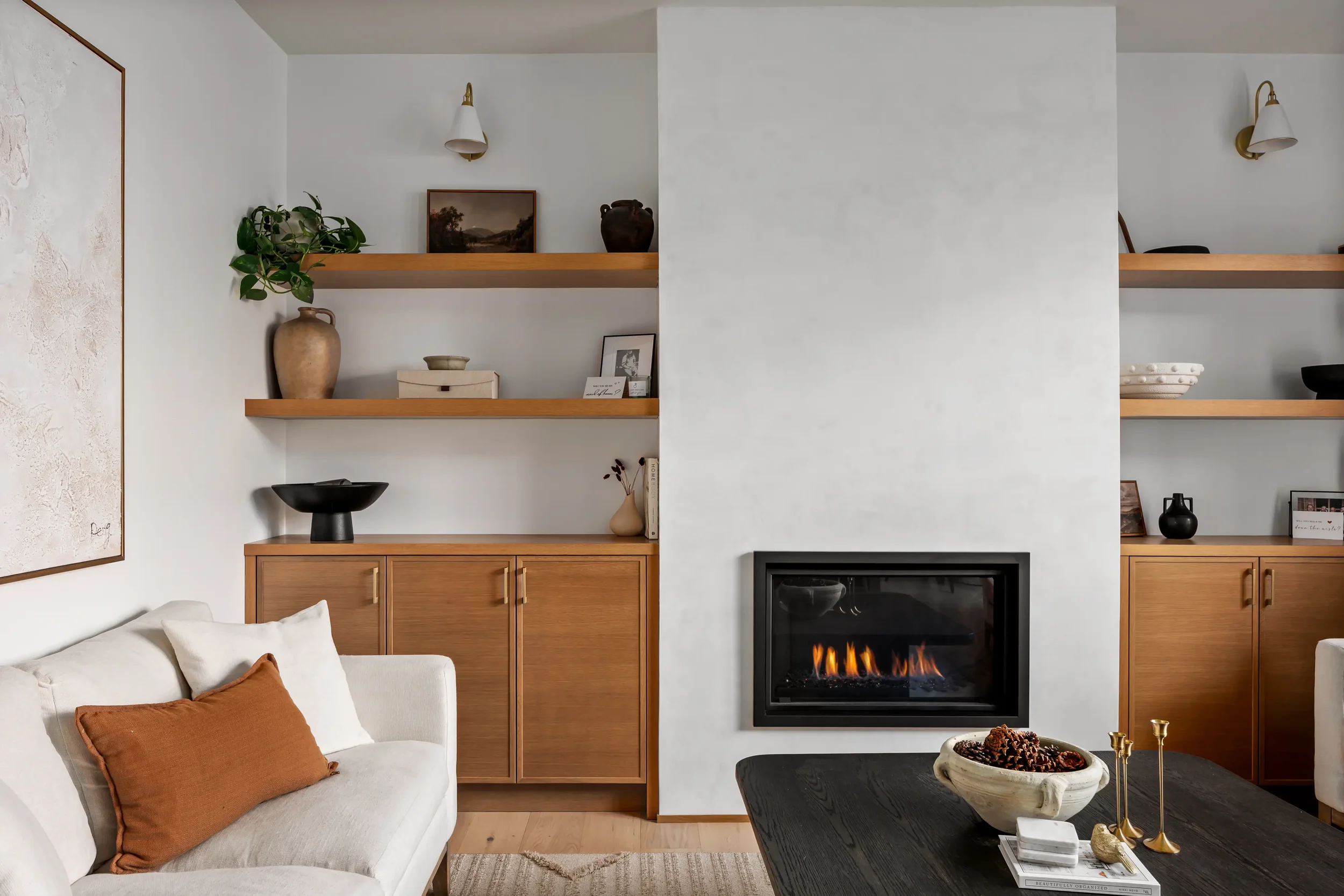 Living room with a white sofa, orange pillows, a black coffee table with a large ceramic bowl of pinecones, a fireplace, and wooden shelves with decorative items. The wall has a large framed map and a painting. There are additional decorations on the shelves, including a plant, vases, and picture frames.