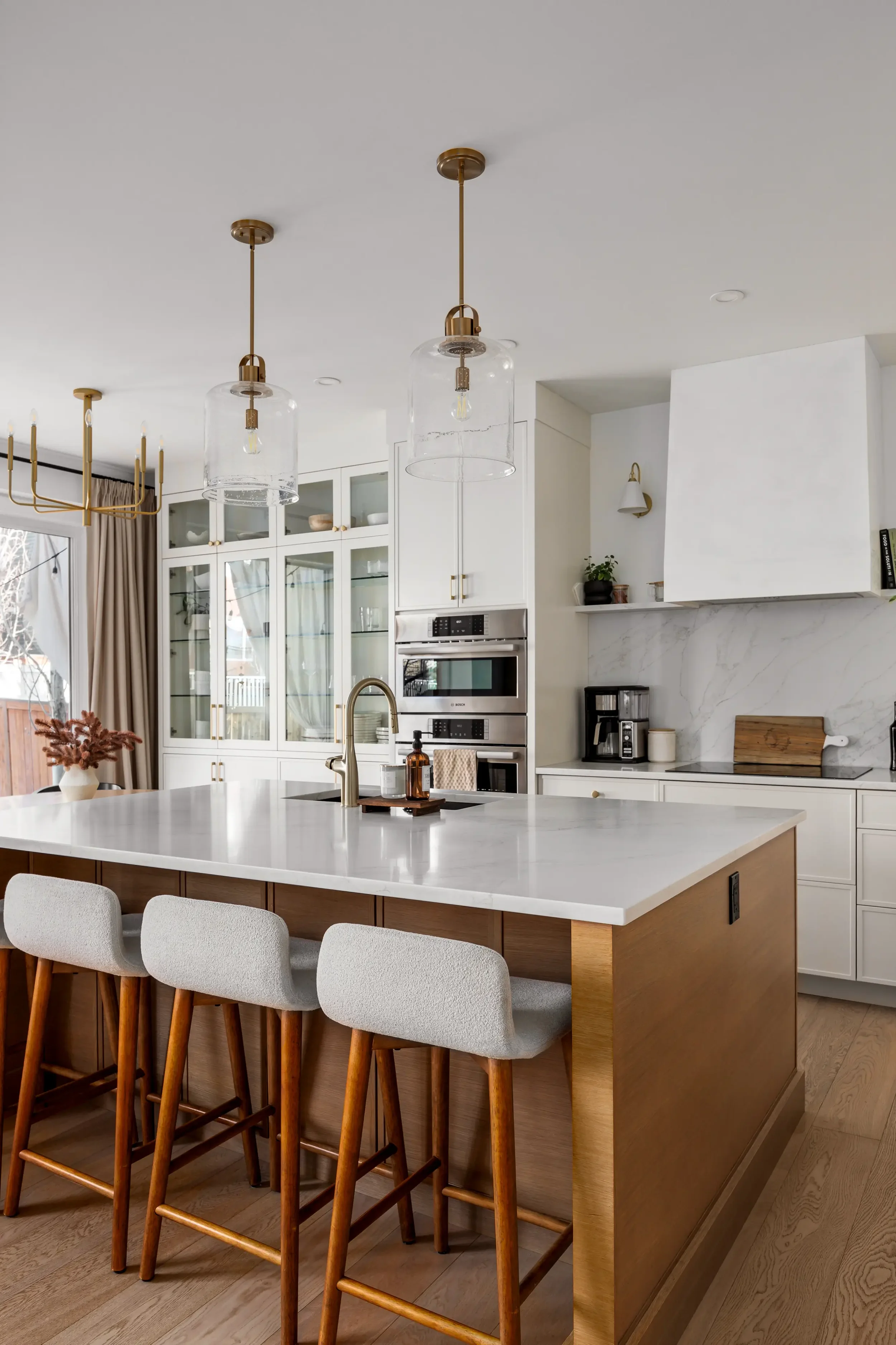 Modern kitchen with a large island with a white countertop, four beige and wooden barstools, pendant lights, white cabinetry, and built-in appliances.