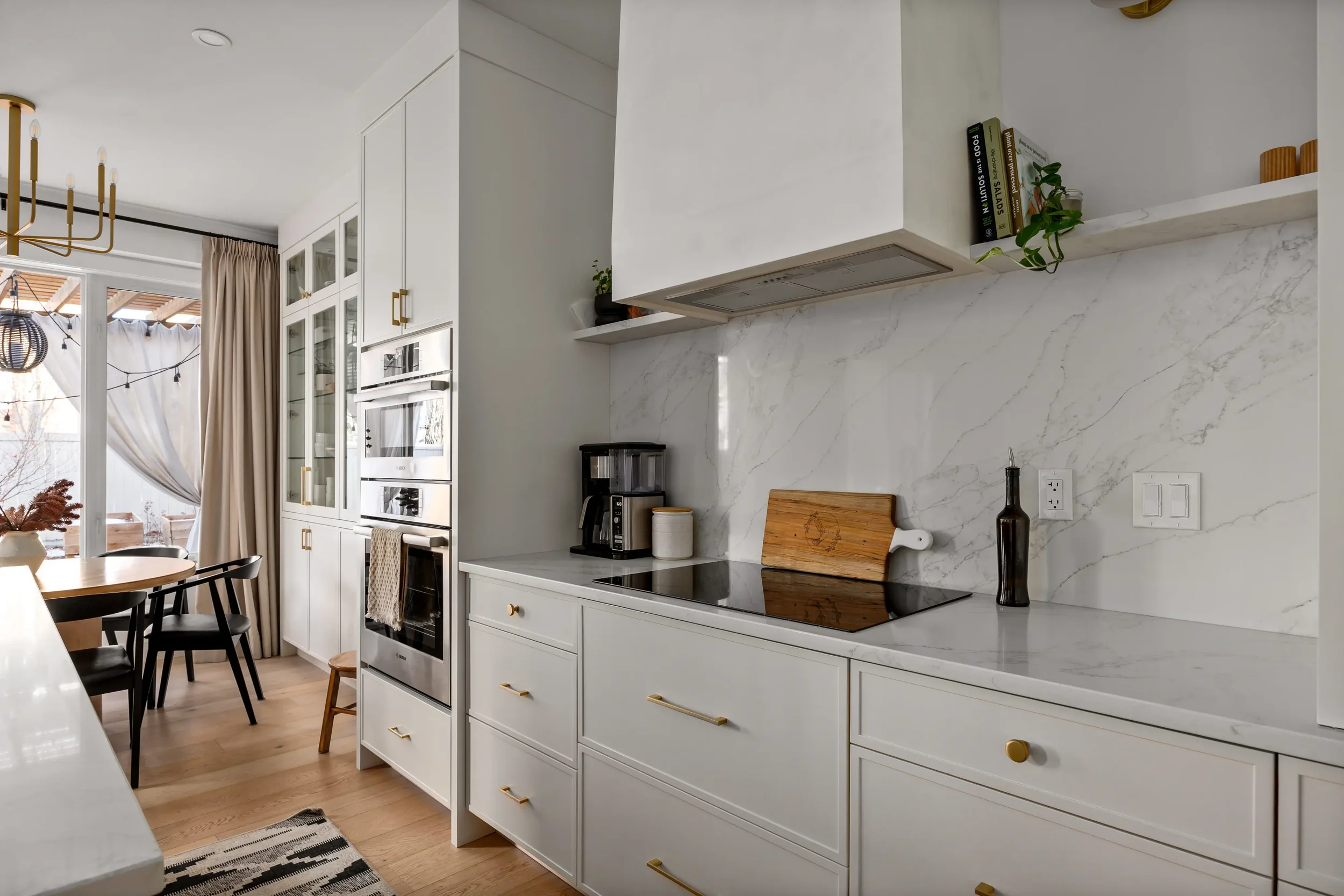 Modern kitchen with white cabinetry, marble backsplash, and open shelving with books and plants. There is a black stovetop, a coffee machine, and decorative items on the counter. In the background, a dining area with a round table and black chairs is