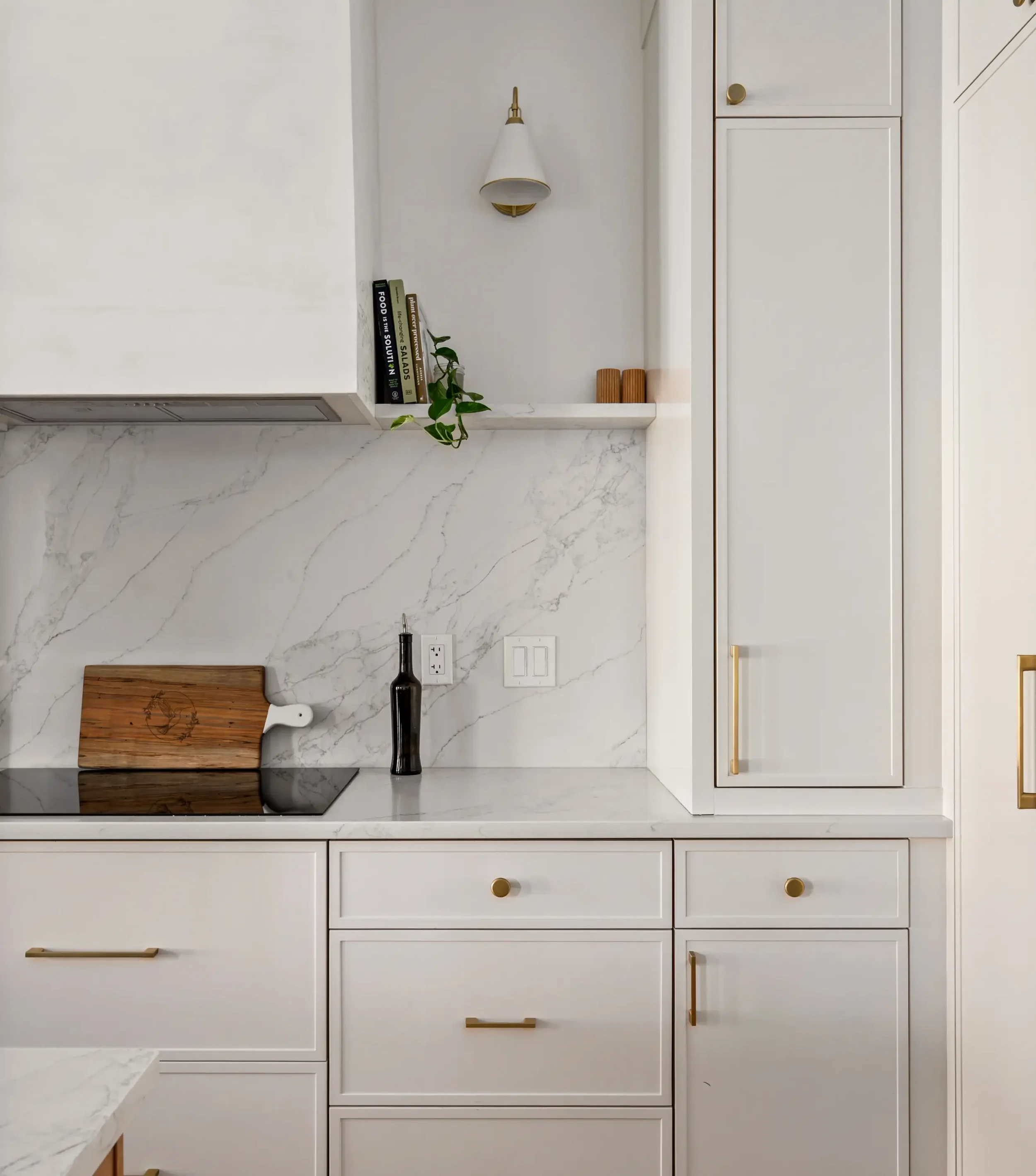 White kitchen with marble countertops, white cabinets with gold handles, a cutting board with a white chef's knife, a black bottle, a small potted plant, books, and a white wall with electrical outlets and switches.