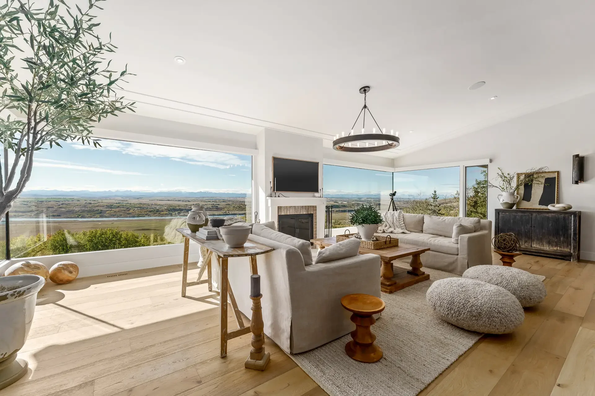 Living room with large windows showcasing mountain and river views, white couches, a wooden coffee table, potted plants, decorative objects, and a black fireplace.