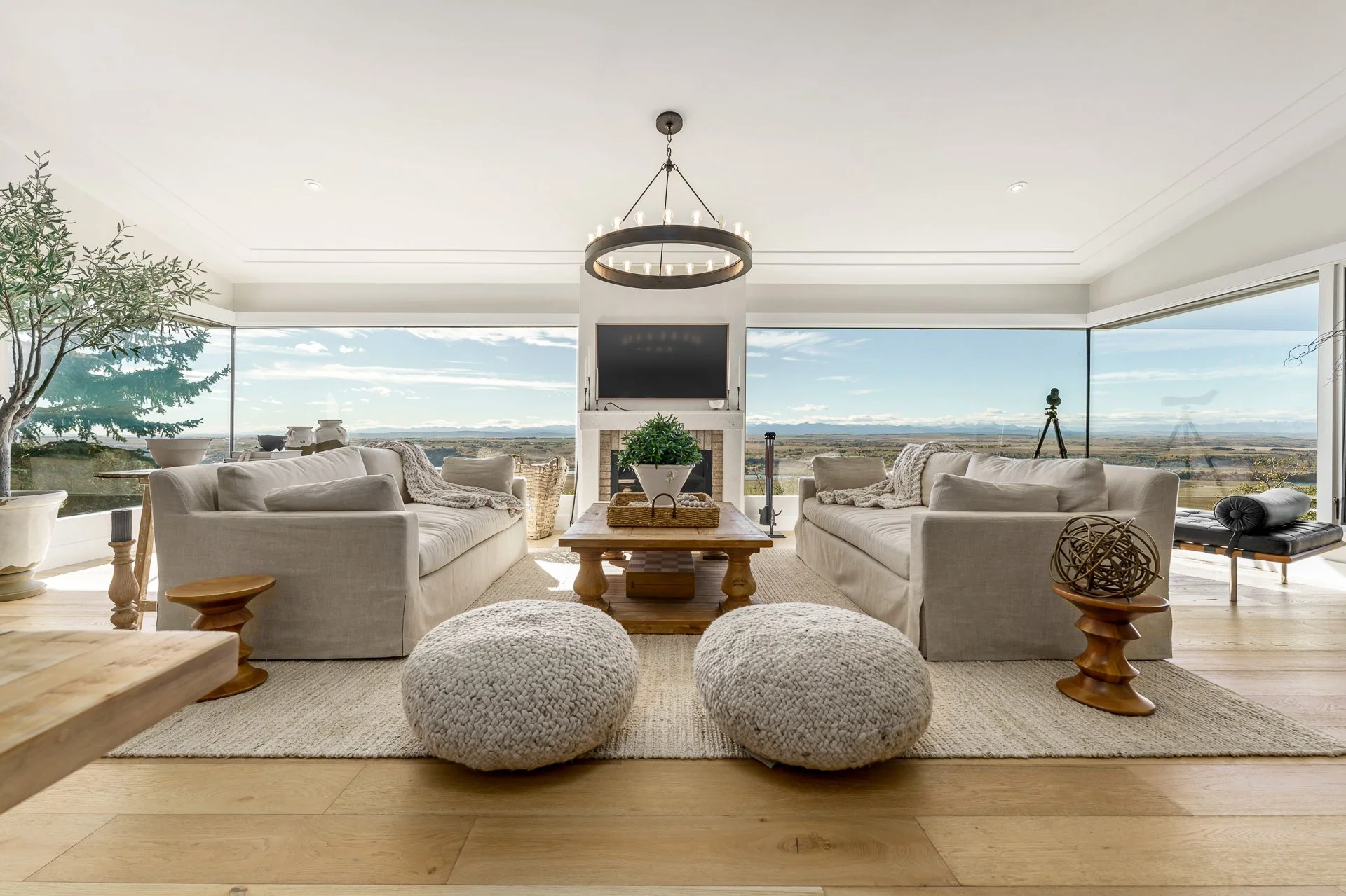 Bright living room with large windows overlooking a landscape, featuring beige sofas, a wooden coffee table, decorative poufs, and a fireplace with a TV above, natural light, and minimalistic decor.