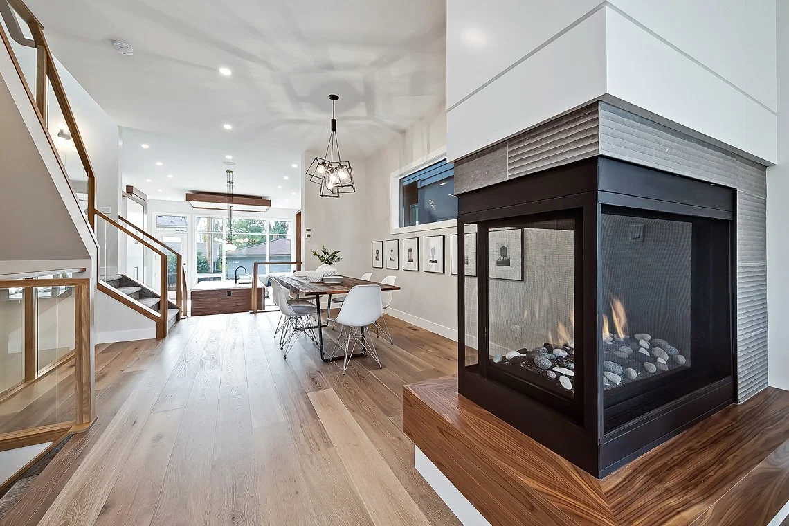 Modern living room with a double-sided fireplace, wooden flooring, dining table with white chairs, staircase, and large windowed door leading outside.