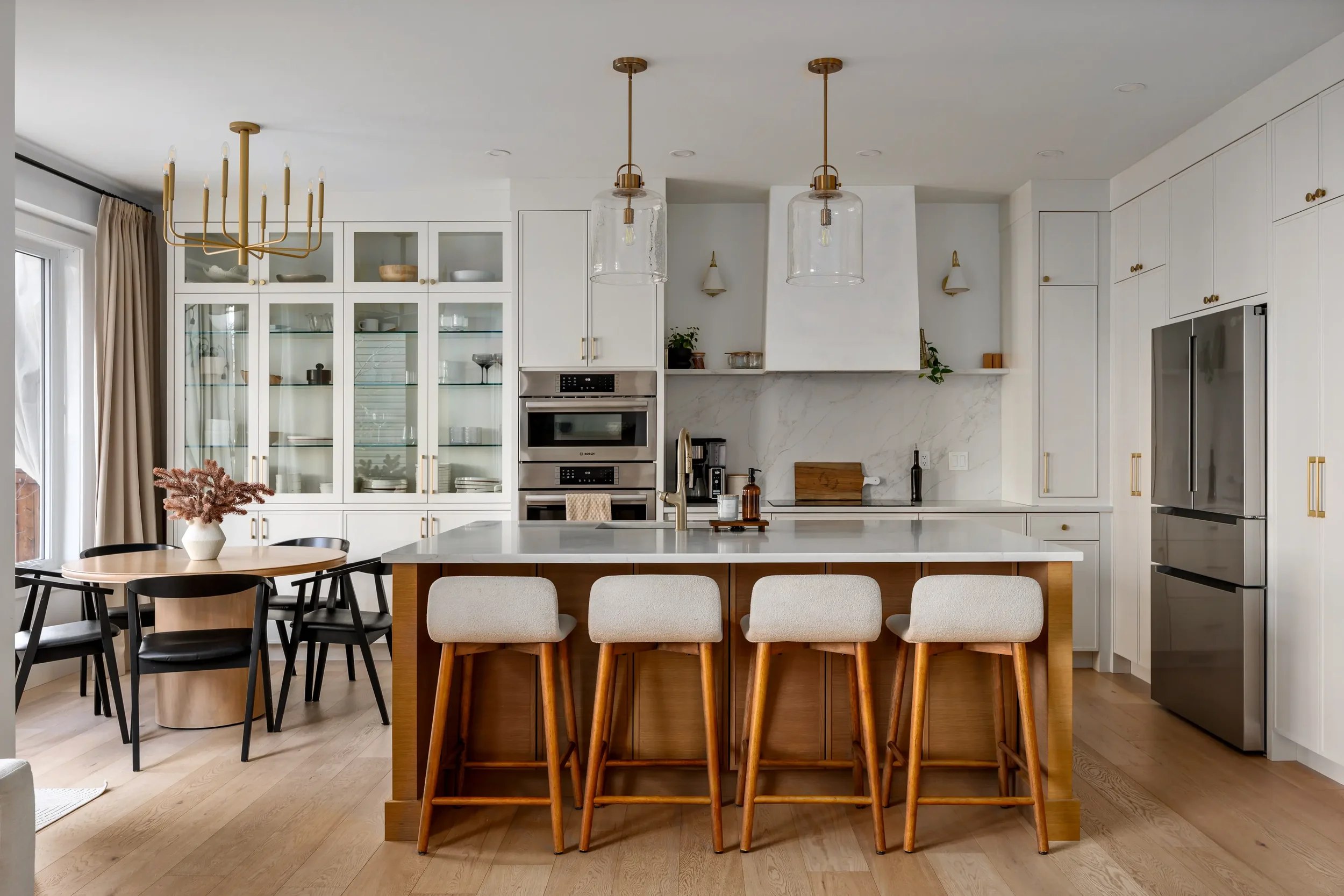 Modern kitchen with white cabinets, a large kitchen island with wooden bar stools, glass pendant lights, and a glass display cabinet. There is a dining table with black chairs on the left and hardwood flooring throughout.