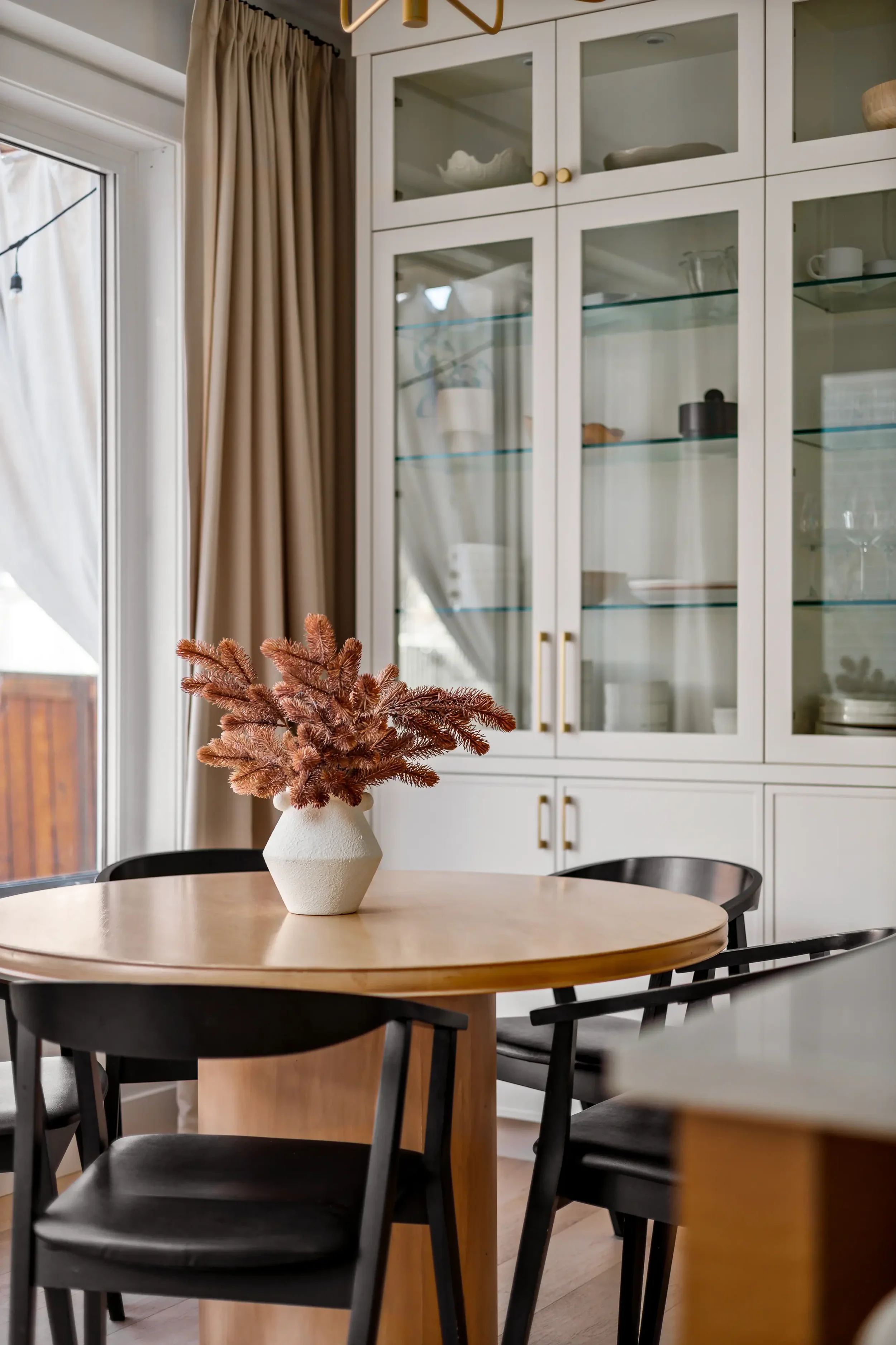 A dining room with a round wooden table, black chairs, a white vase with dried pink flowers, a large white cabinet with glass doors, beige curtains, and a sliding glass door to the outside.