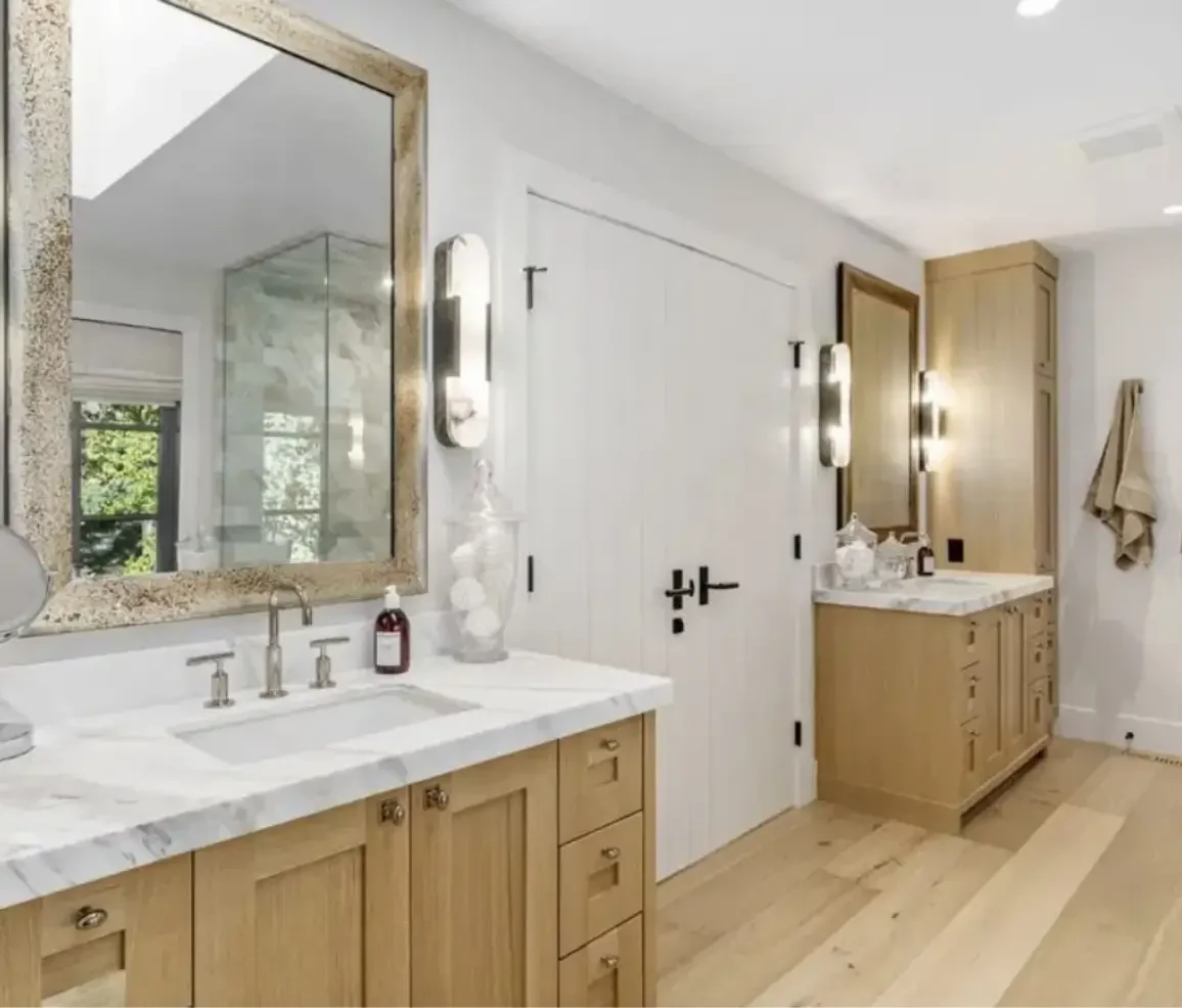 A luxurious bathroom with two wooden vanities, each with a marble countertop, mirrors, and modern light fixtures. There is a window with a view of greenery, and the room has light-colored wooden flooring and white walls.
