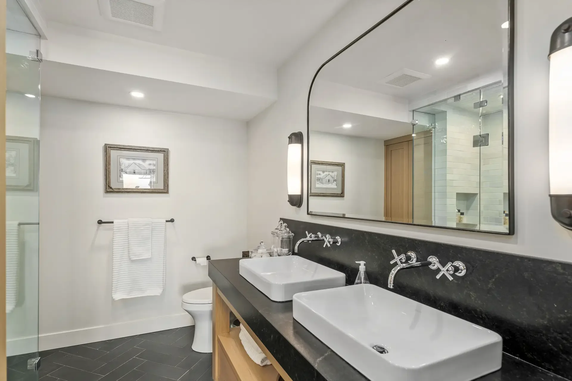 Modern bathroom with double vessel sinks on black countertop, large mirror, wall-mounted lights, framed art, towel rack, and glass shower enclosure.