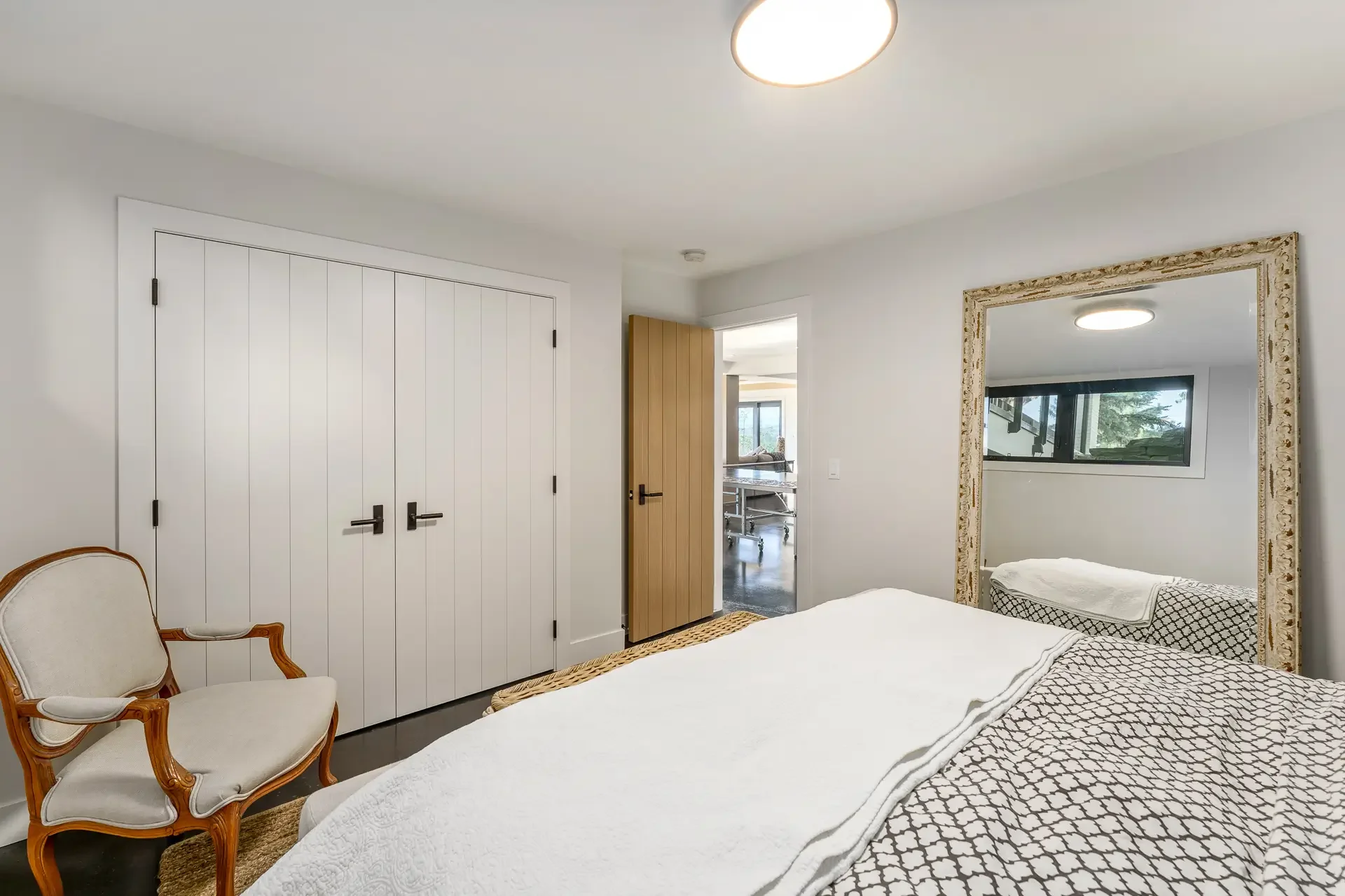 A bedroom with a large mirror on the wall, a white bed with patterned bedding, a wooden chair with a white cushion, white double doors, an open door to another room, and a ceiling light.