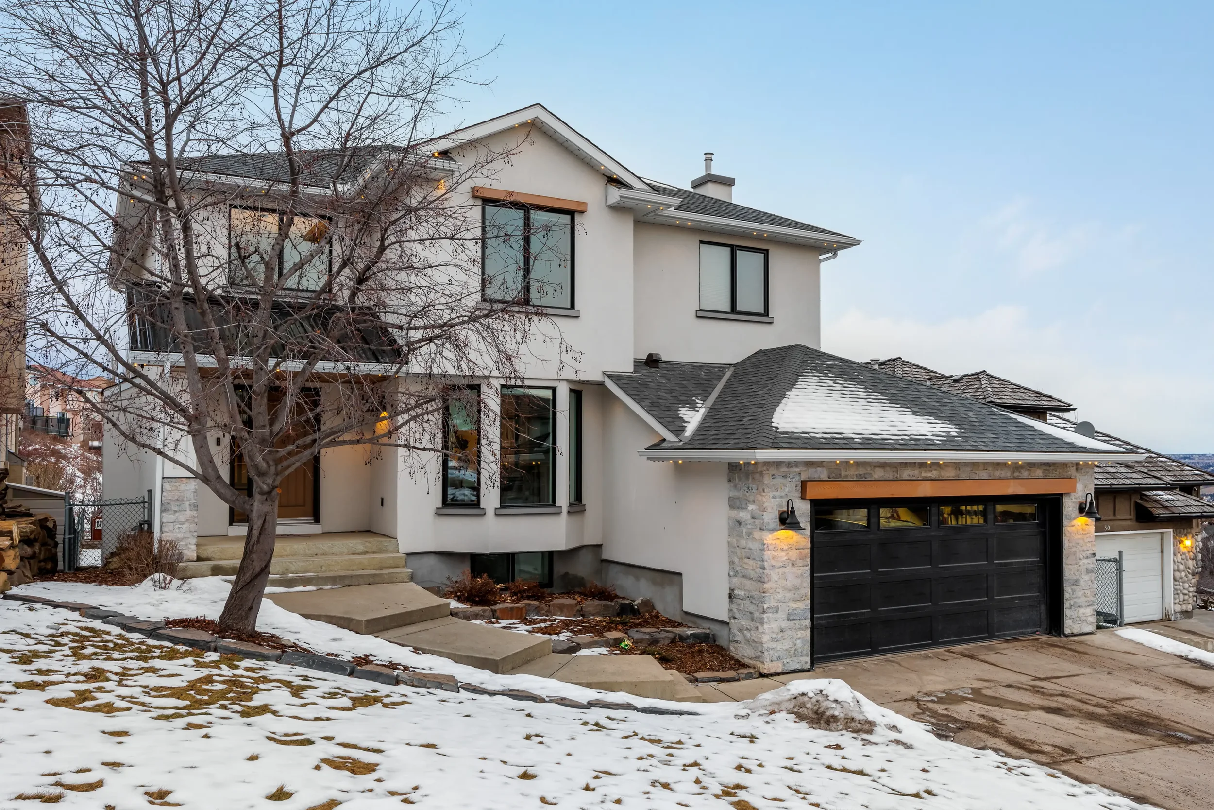 Modern multi-story house with large windows, a gray garage door, snow on the ground, and a leafless tree in front.