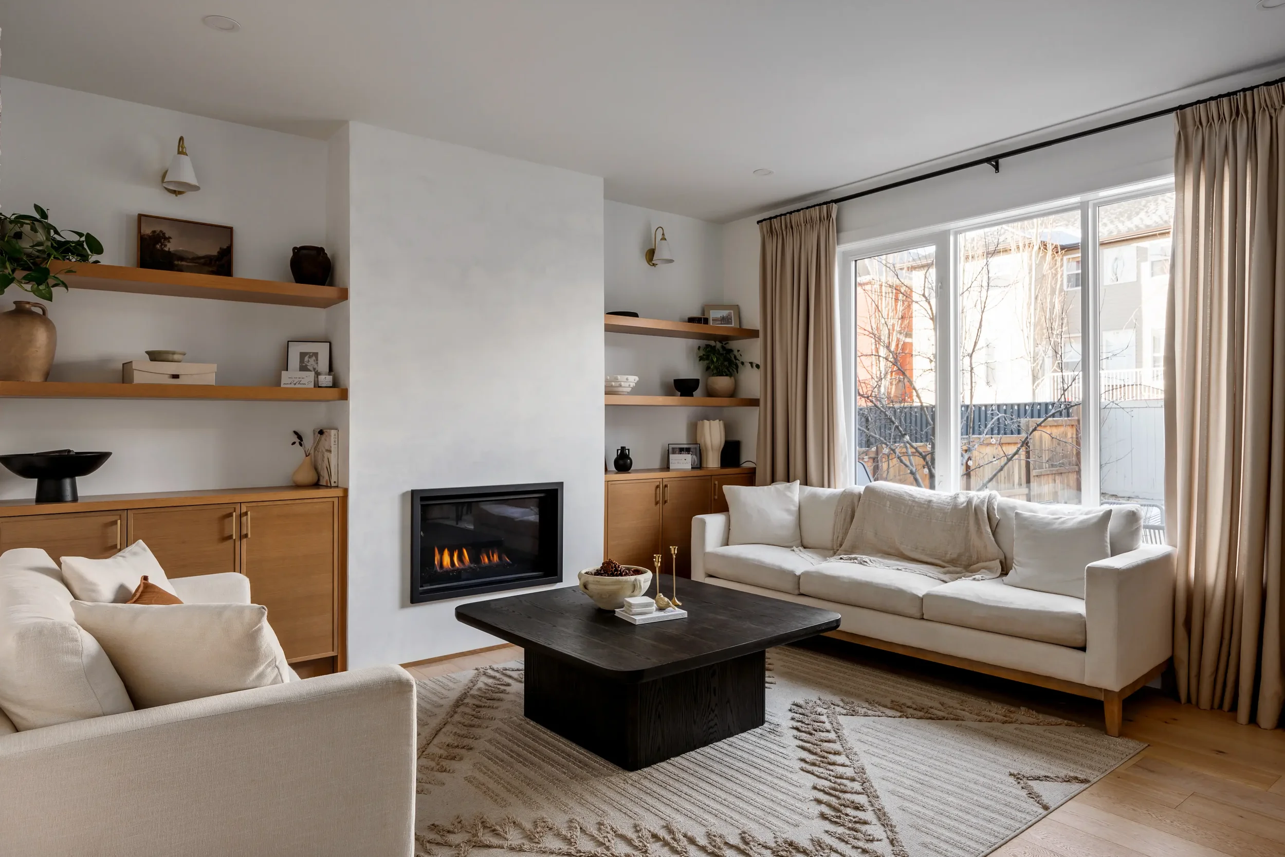 A cozy living room with white sofas, a dark wooden coffee table, and a fireplace. Large windows with beige curtains let in natural light, showing a backyard with trees. Shelves with decorative items and a TV are also visible.