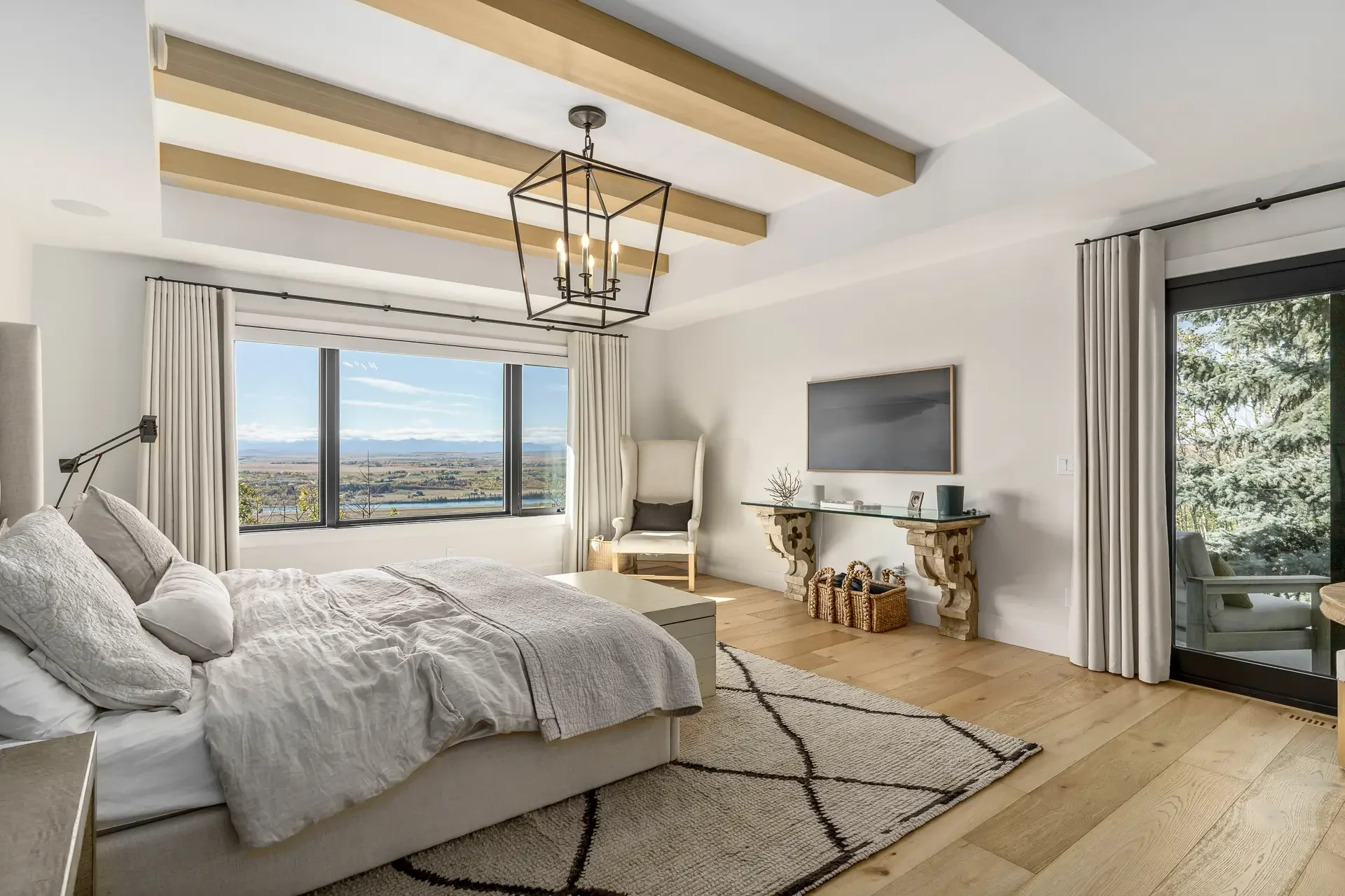 A cozy bedroom with large windows offering a scenic view of a distant landscape, featuring a bed with white linens, a modern chandelier, a metro map wall decor, and sliding glass doors with white curtains.