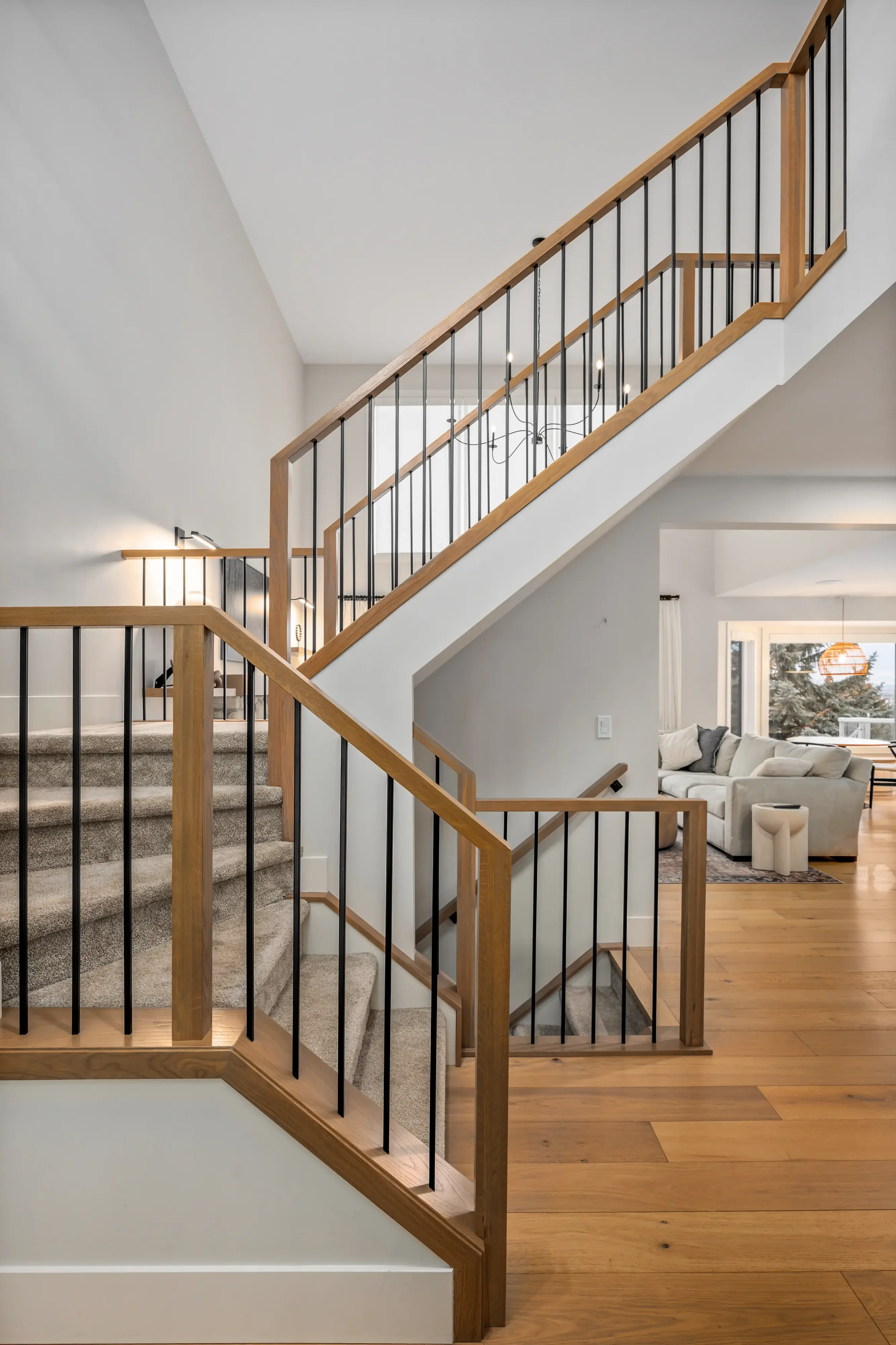 Interior view of a staircase with beige carpeted steps and a wooden and black metal railing, leading to the second floor of a home, with a living room visible in the background.