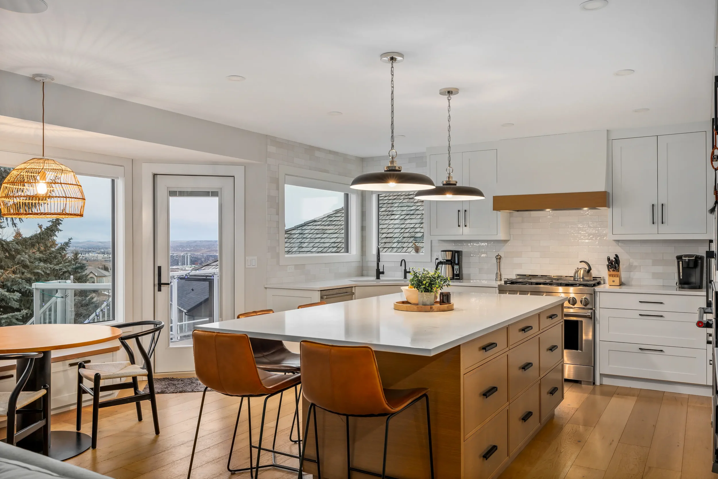 Modern kitchen with white cabinets, stainless steel oven, island with wooden drawers, black and brown chairs, pendant and woven pendant lights, windows showing a cityscape and mountains, potted plant on island.