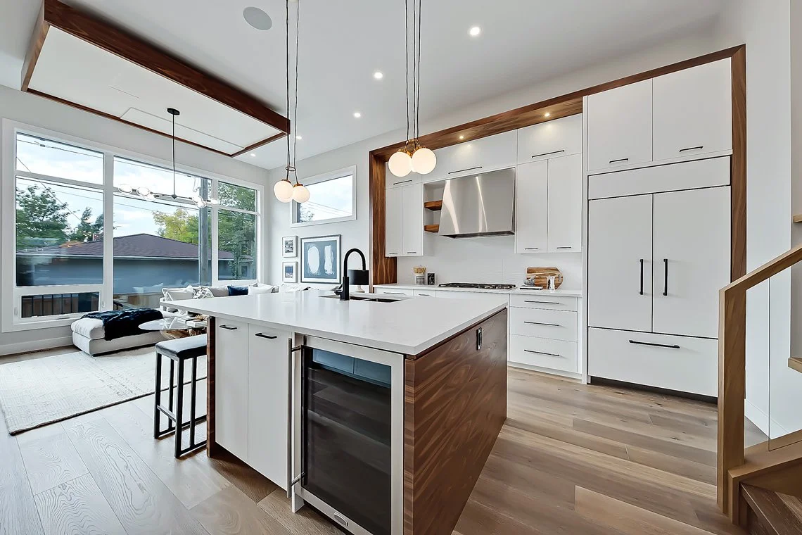 Modern kitchen with white cabinets, a large island with a built-in wine fridge, and a living area with large windows and a white sofa.