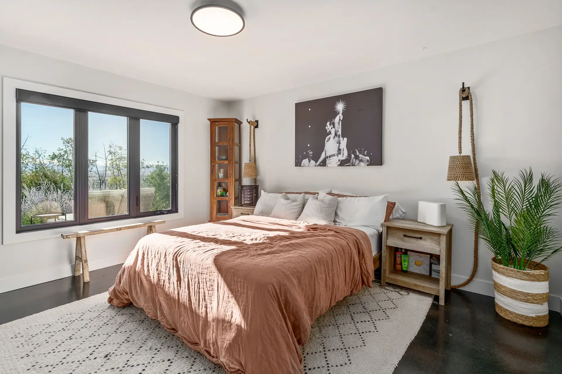 A bedroom with a large bed covered with a brownish-orange blanket and multiple pillows. There is a window to the left with a black frame showing an outdoor view of trees and a patio. Beside the bed, there is a small wooden side table with a few items