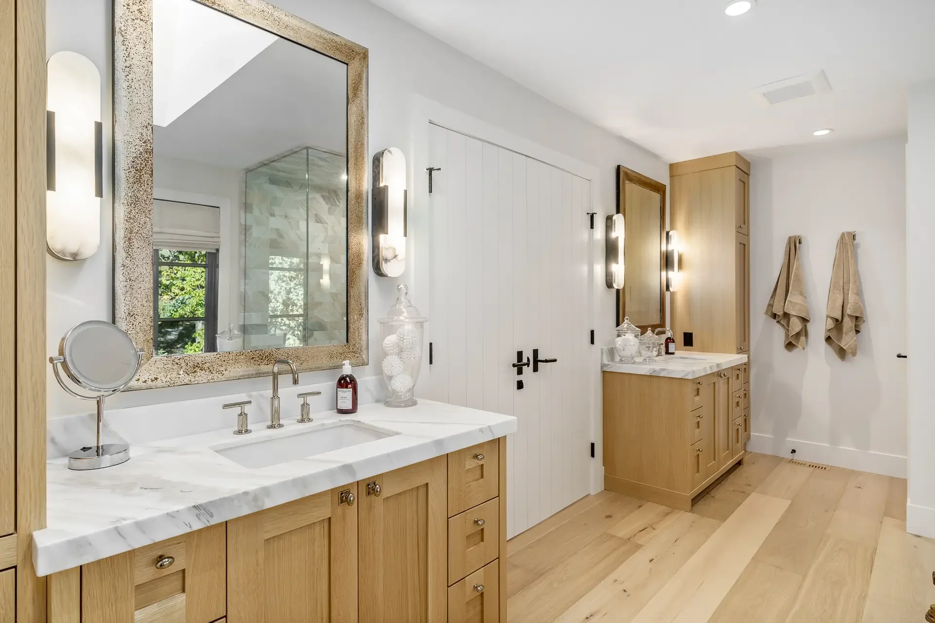 A modern bathroom features a white marble vanity with a built-in sink, wooden cabinets, and a large mirror with a textured gold frame. There are two wall-mounted light fixtures beside the mirror, a small round magnifying mirror, and decorative items 