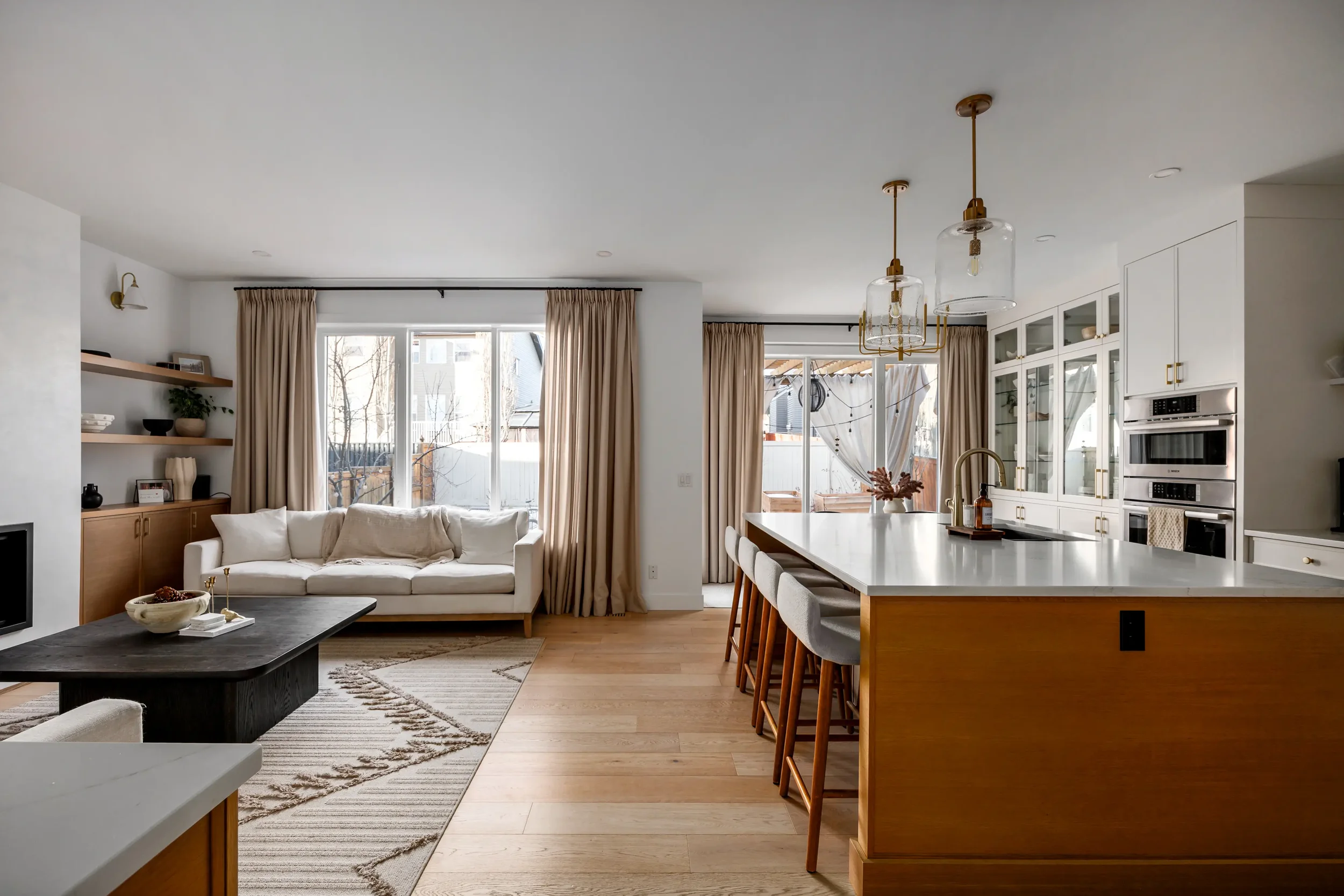 Bright living room and kitchen area with large windows, beige curtains, white cabinetry, and a kitchen island with bar stools.