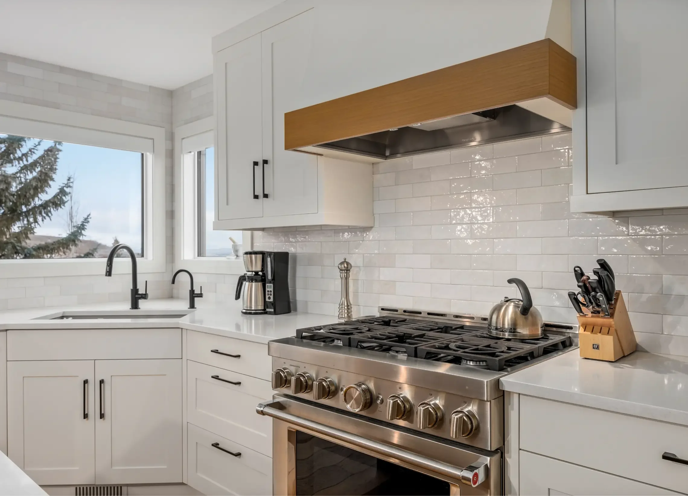 Modern kitchen with white cabinets, stainless steel stove, and a white tiled backsplash. There are two windows above the sink, with trees visible outside. On the countertop, there is a coffee maker, a kettle, and a knife block.