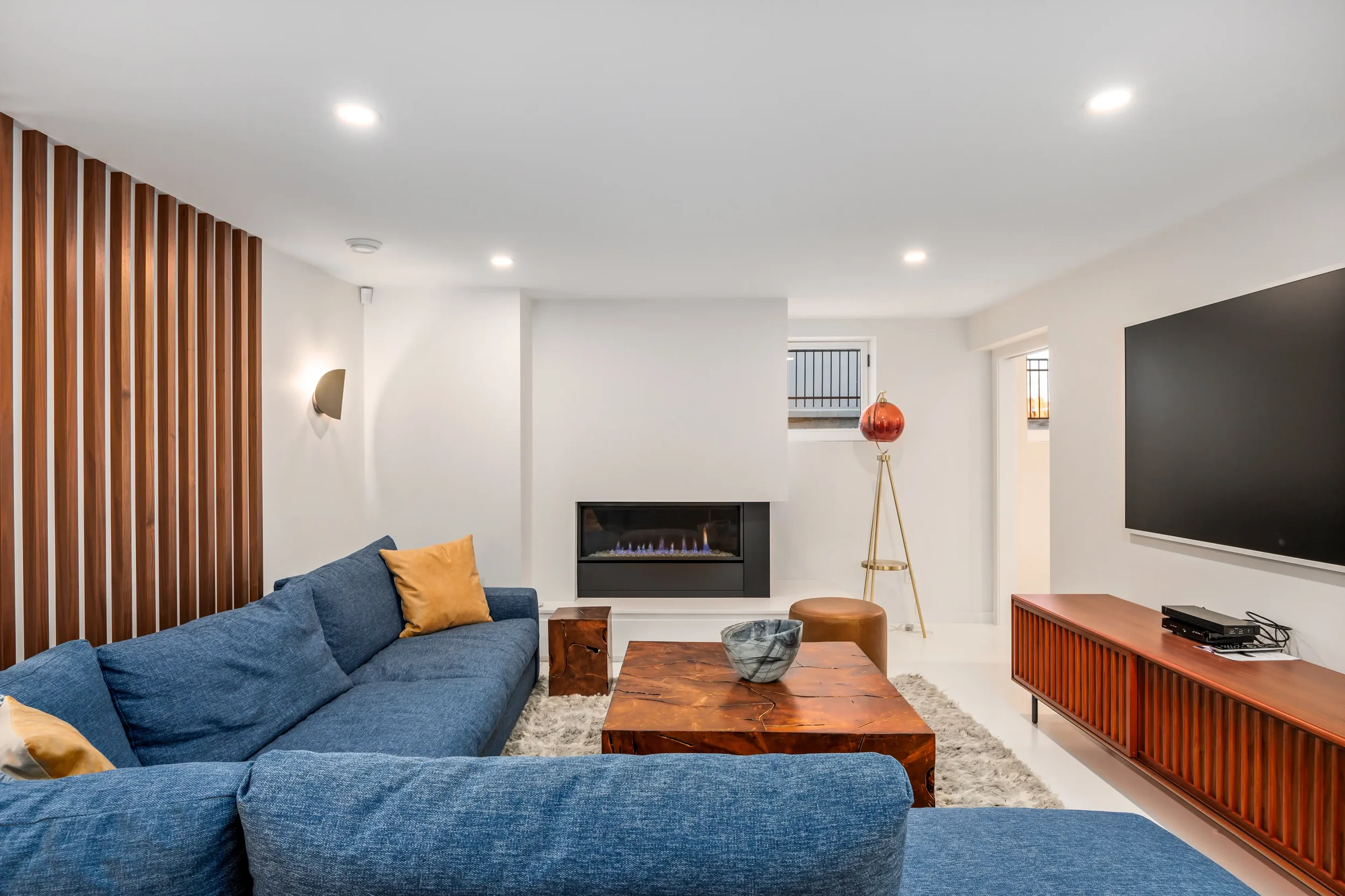 Living room with blue sectional sofa, wooden coffee table, gas fireplace, wall-mounted TV, decorative lamp, and wooden slat accent wall.