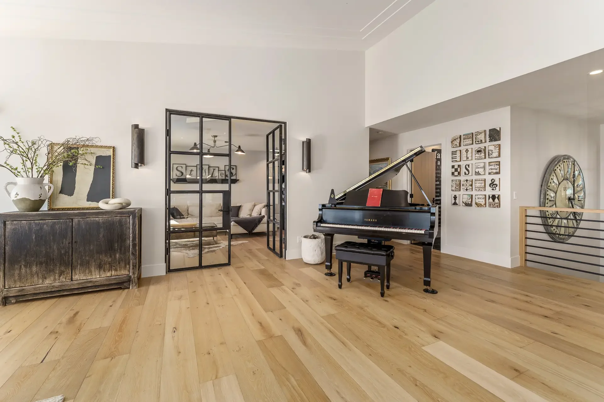Modern living room with a Yamaha grand piano, a wooden sideboard with a large vase of greenery, and artwork on the walls. French doors lead to a sitting area with a sofa, and the room features hardwood floors and decorative elements.