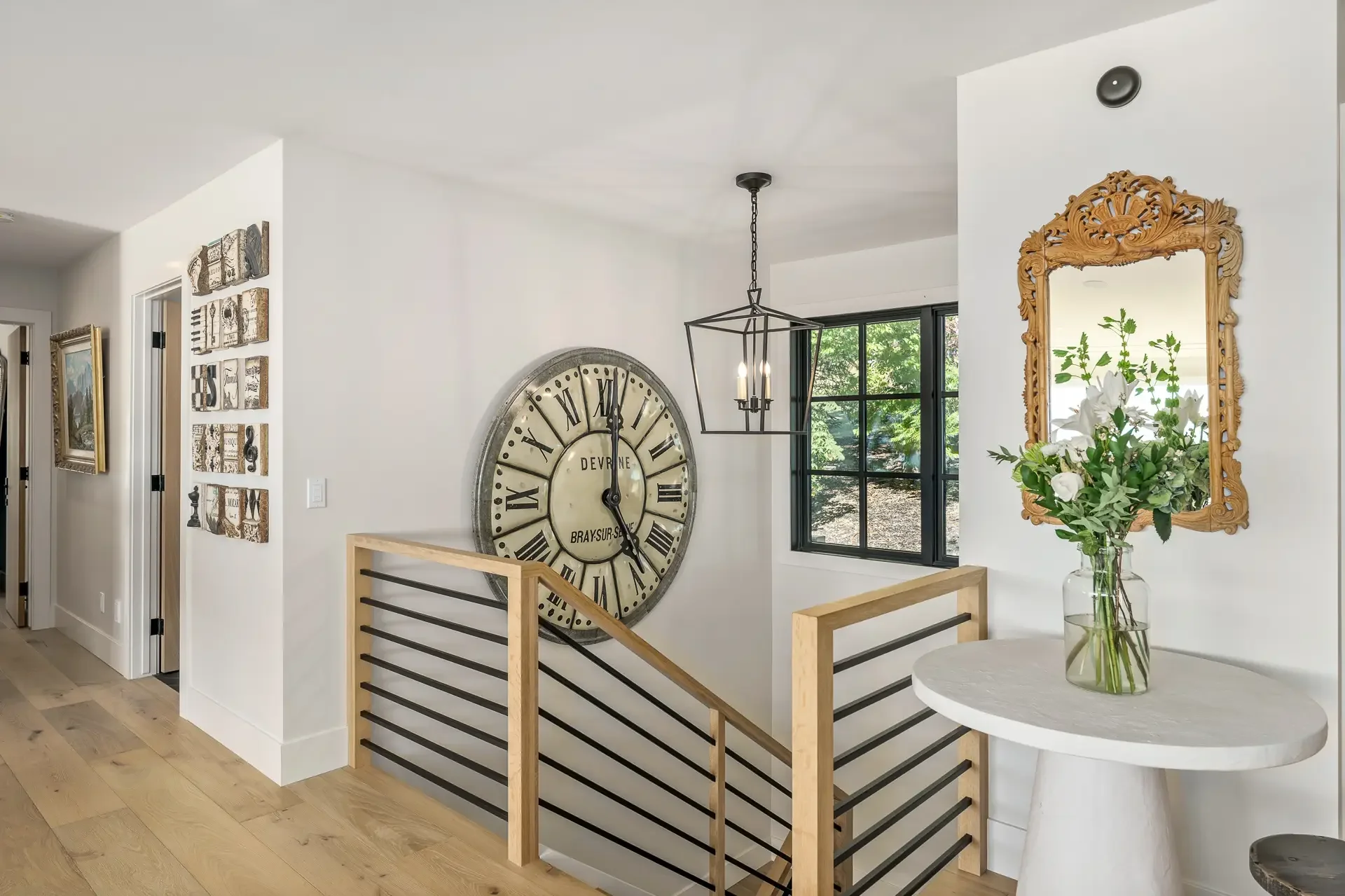 Interior view of a modern house stairwell with a large vintage clock on the wall, a geometric chandelier, a window showing greenery outside, a decorative mirror with a wooden frame, and a table with a vase of white flowers.