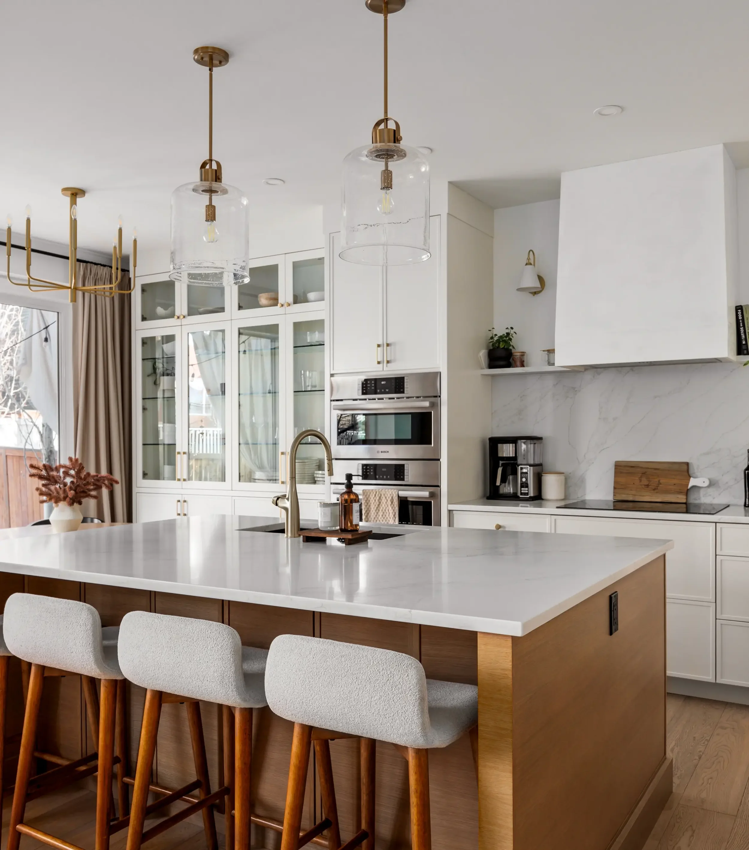 Modern kitchen with white cabinets, a large white marble island with a gold sink, and bar stools with white cushions and wooden legs. Overhead, two glass pendant lights hang from a gold fixture. In the background, a gold chandelier with candles and a tree branch decoration are visible near a window with curtains. The kitchen features built-in stainless steel appliances and a marble backsplash.