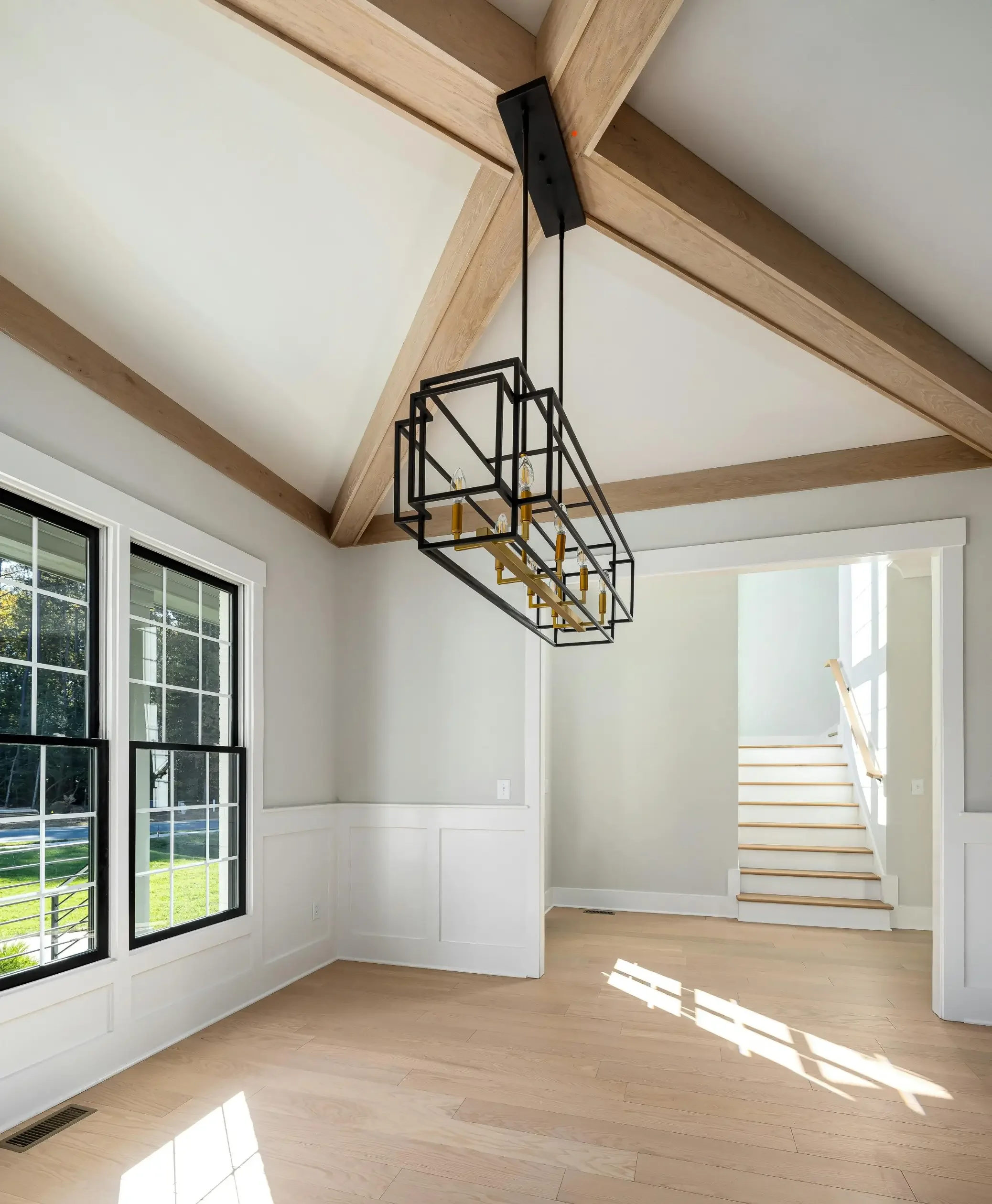 Image of an empty living room with large windows, white walls, and light wooden floors. A staircase with light wood steps and a staircase handrail leads up to the second floor. A black chandelier with multiple geometric frames and exposed light bulbs hangs from the ceiling.