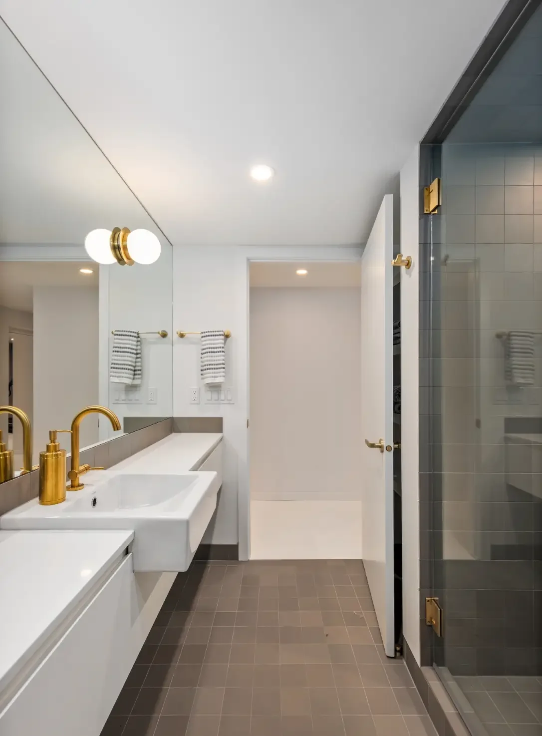 Modern bathroom with white vanity, gold fixtures, large mirror, striped towels, gray tiled shower, and neutral floor tiles.