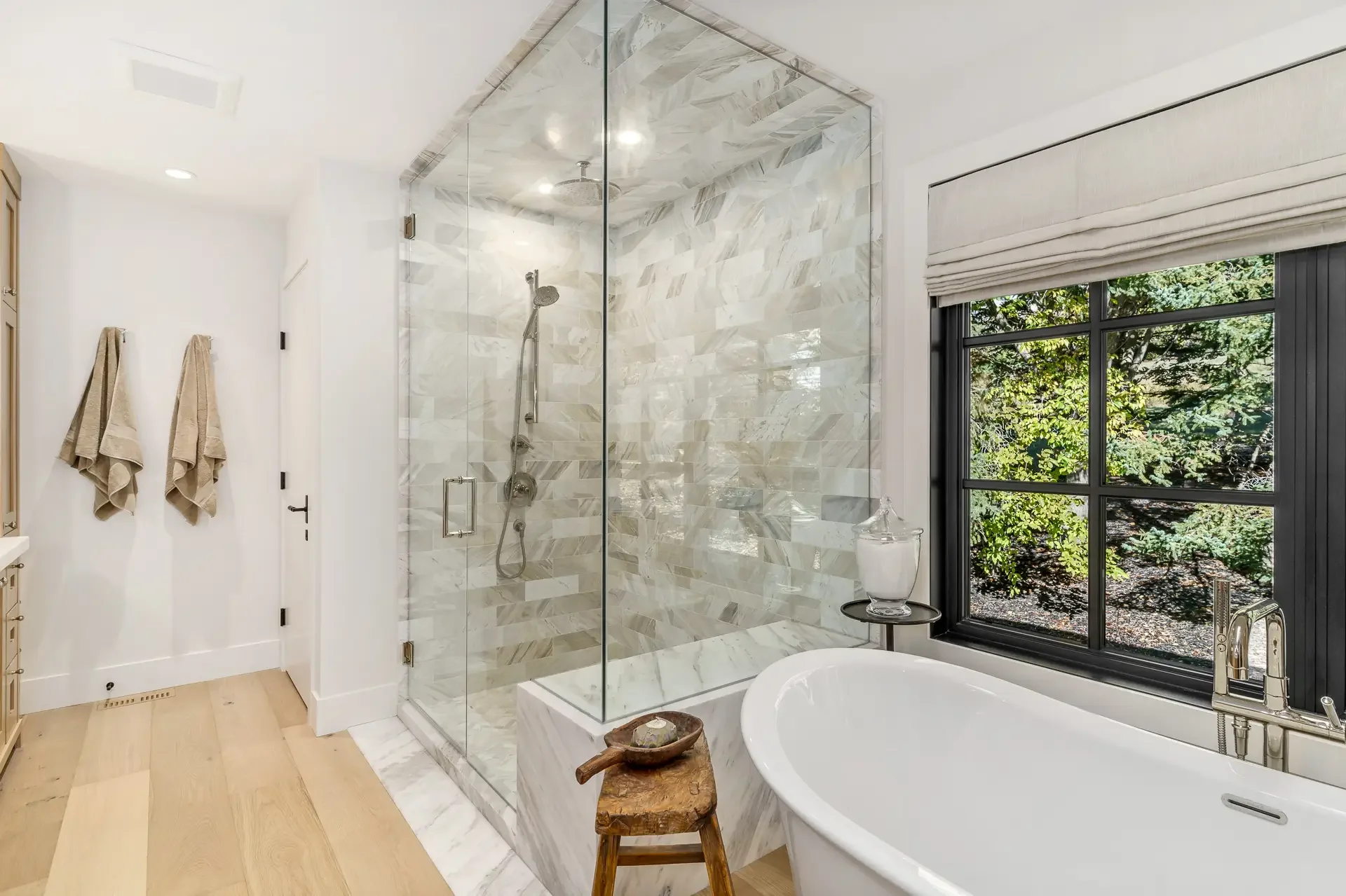 Bathroom with a walk-in shower featuring marble tiles, a large window showing greenery outside, and a bathtub with a faucet, a small wooden stool, and beige towels hanging on the wall.