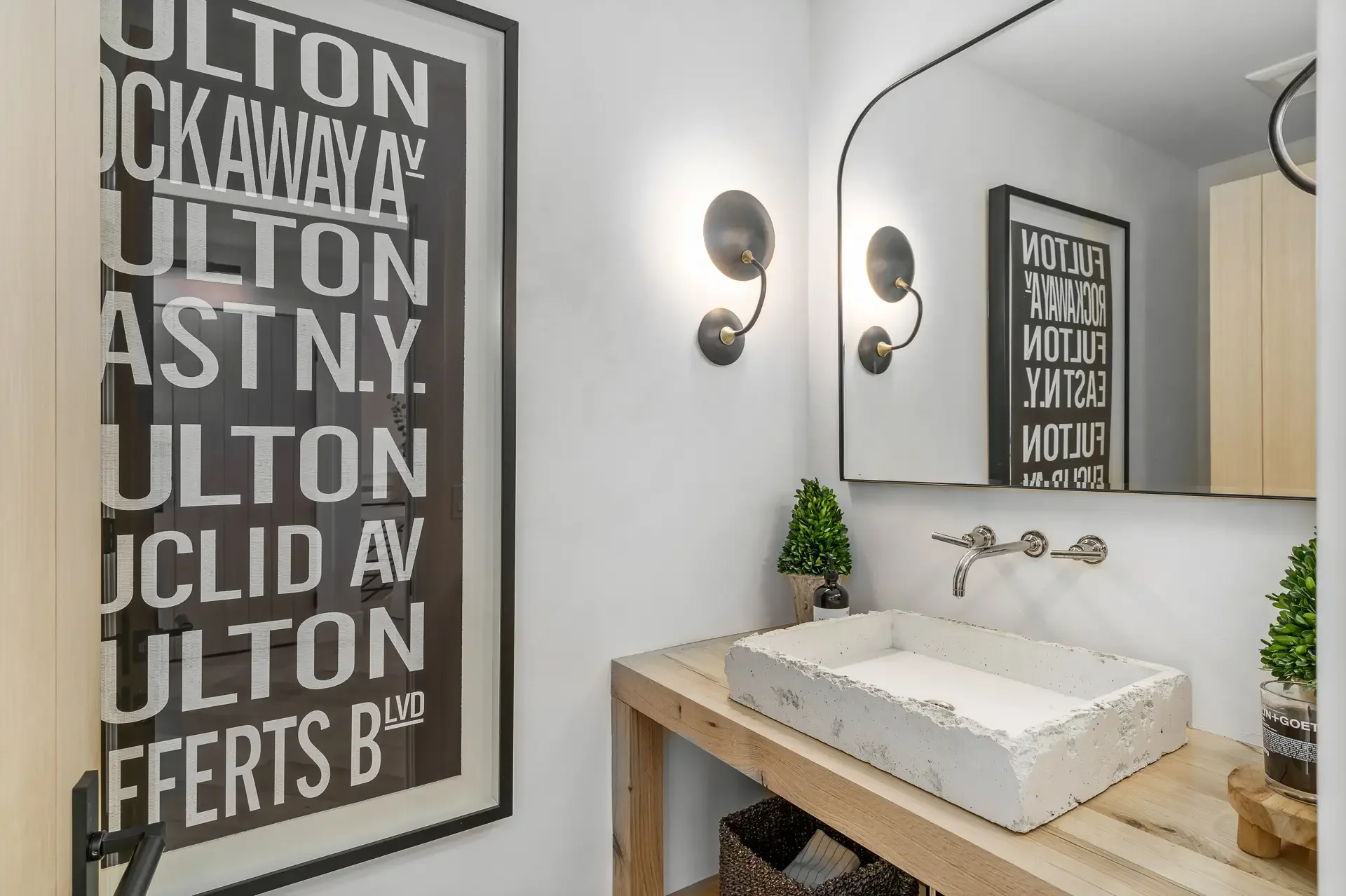 Modern bathroom featuring a large framed mirror above a concrete sink with wall-mounted faucet, two black wall sconces with brass accents, a small potted plant, and a black and white subway art print on the wall, with natural light illuminating the s