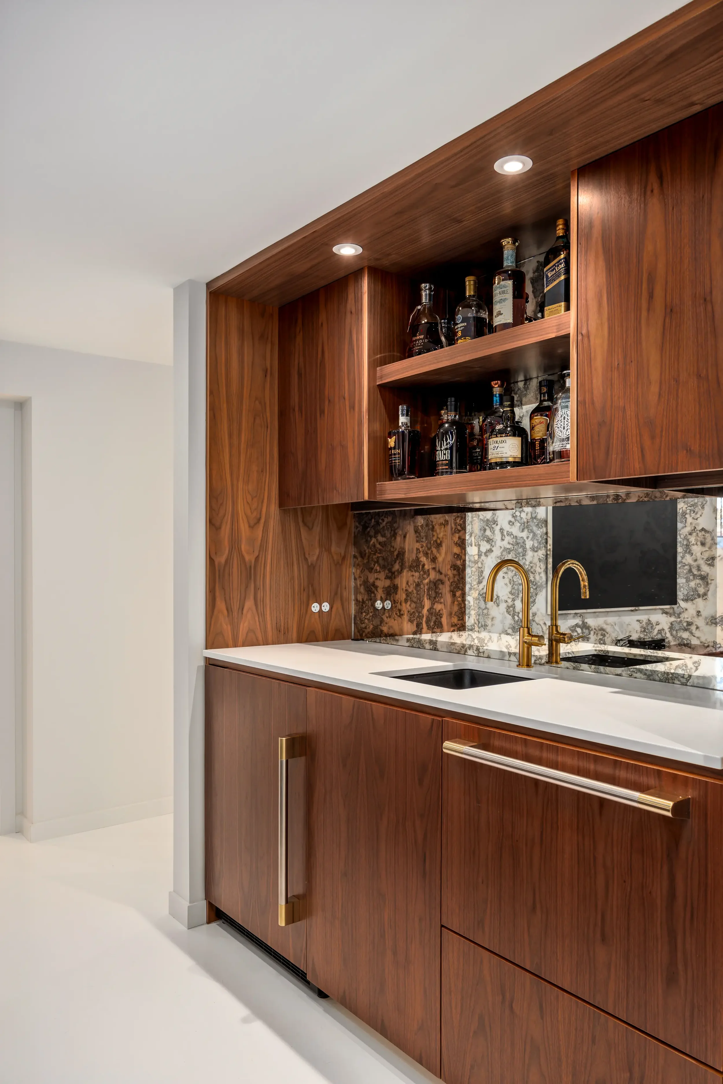 Modern kitchen with wooden cabinets, a marble backsplash, a white countertop, gold fixtures, and a built-in collection of liquor bottles on a shelf.